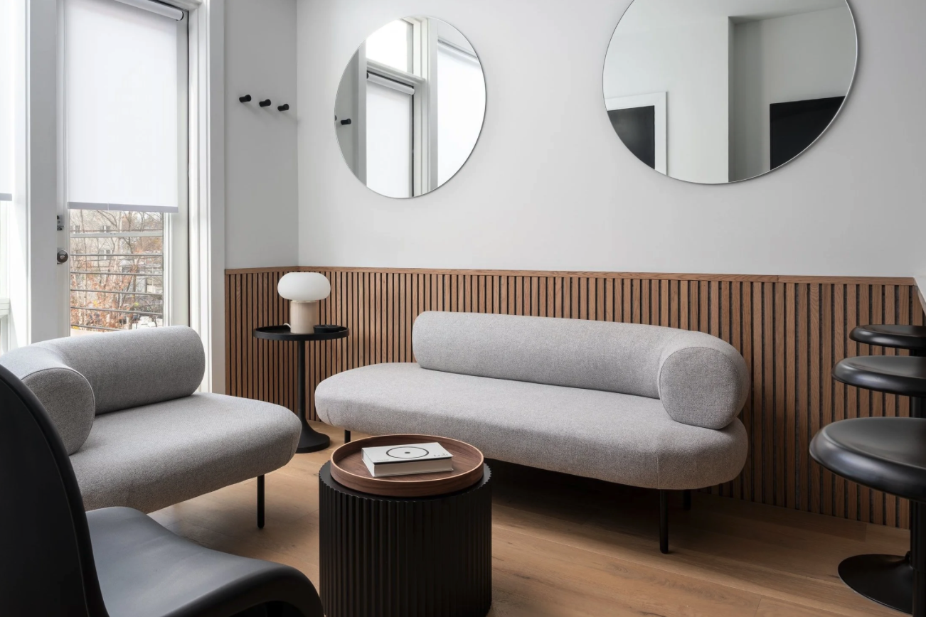 A modern living room with two grey sofas, a wooden side table with a white lamp, a round black coffee table with magazines, and two round mirrors on the white wall. There are windows with white blinds and wooden flooring. Interior design styling