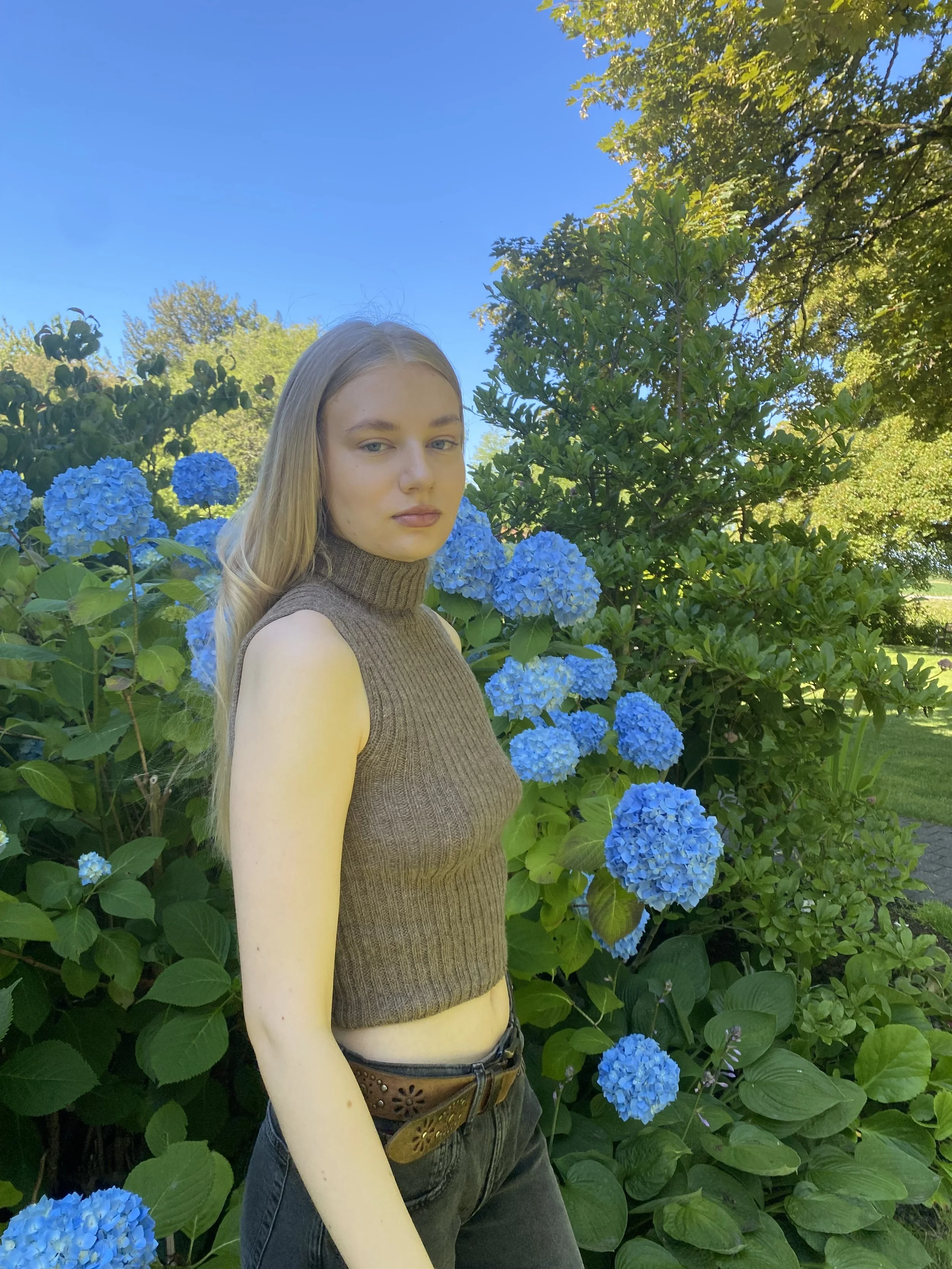 A young woman with long blonde hair and fair skin standing outdoors beside a large bush of blue hydrangea flowers. She is wearing a sleeveless brown turtleneck top and dark jeans, with a brown belt. The background features lush green foliage and a clear blue sky.
