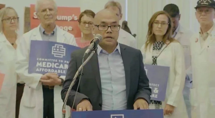 Azlan Ibrahim, public relations and communications professional, speaks at a news conference with healthcare professionals urging protections for Medicare. The doctors are holding signs supporting Medicaid and affordable healthcare in the background.