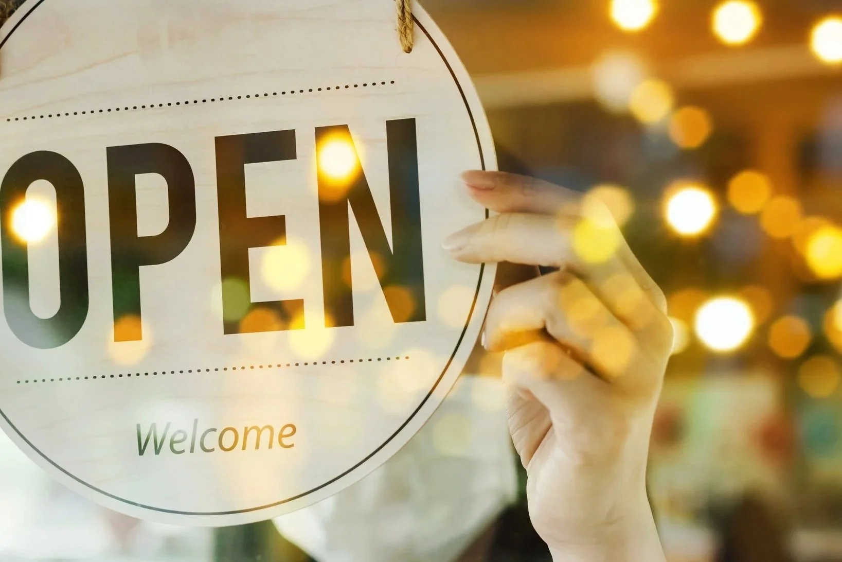 A hand peeking through a glass door holding a circular sign that says 'OPEN' with the word 'Welcome' below it, illuminated with warm golden bokeh lights in the background.