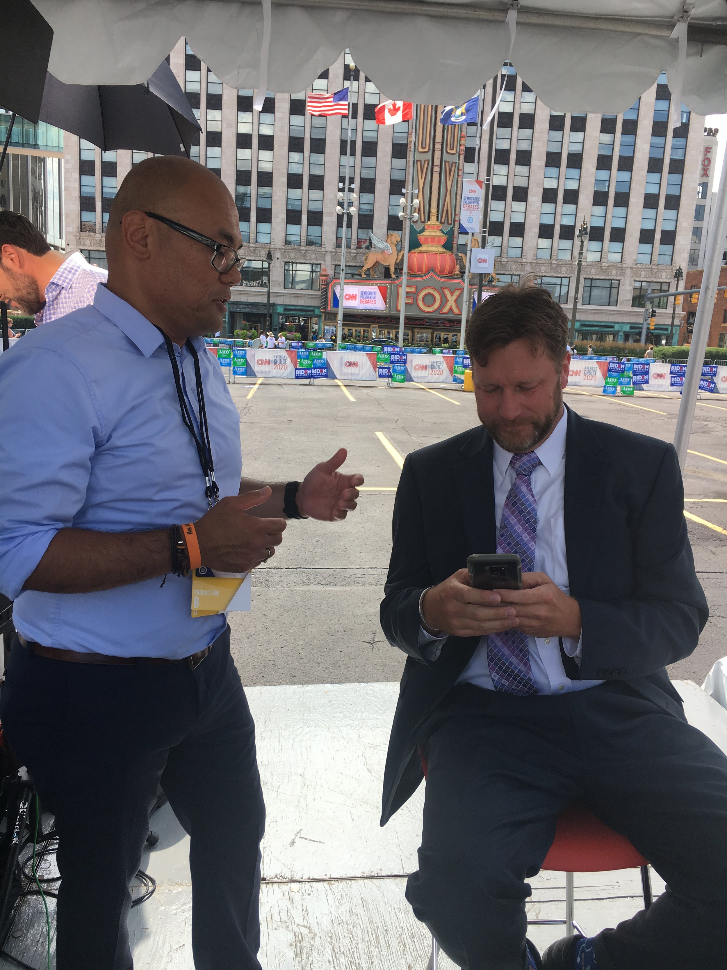 Two men having a conversation outside near the Fox theater, with a large theater sign and flags in the background.