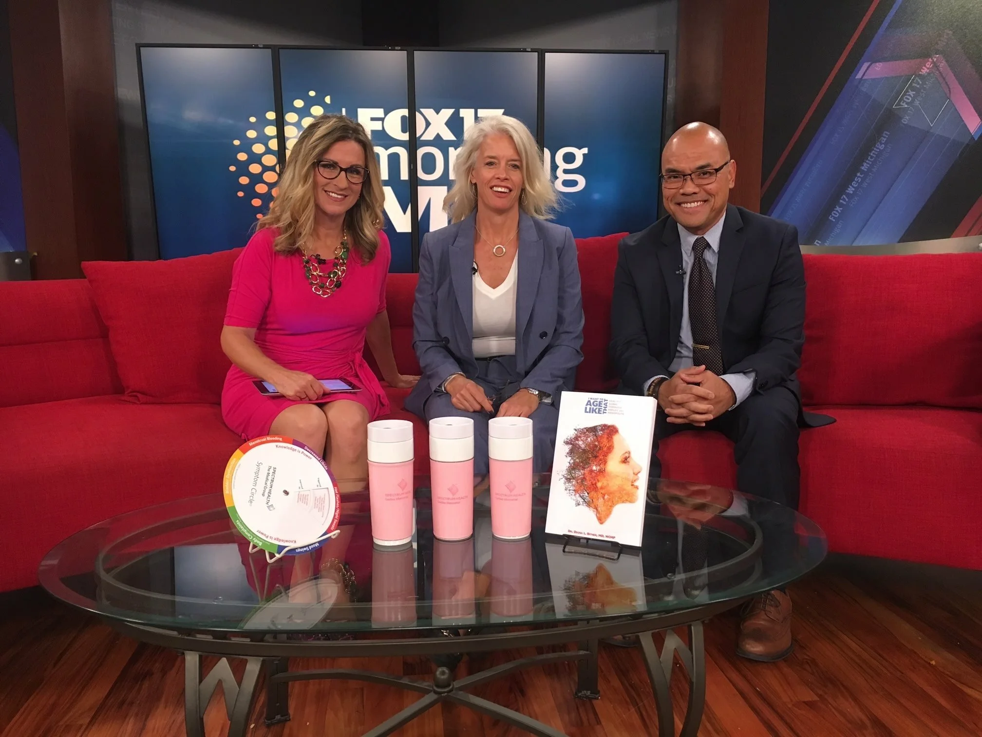 Three people sitting on a red couch in a television studio with 'FOX 17 Morning' logo on screens behind them. There is a glass table with pink products, books, and a white circuit board. The woman on the left is in a pink dress holding a phone, the woman in the middle is in a blue blazer, and the man on the right is in a dark suit.