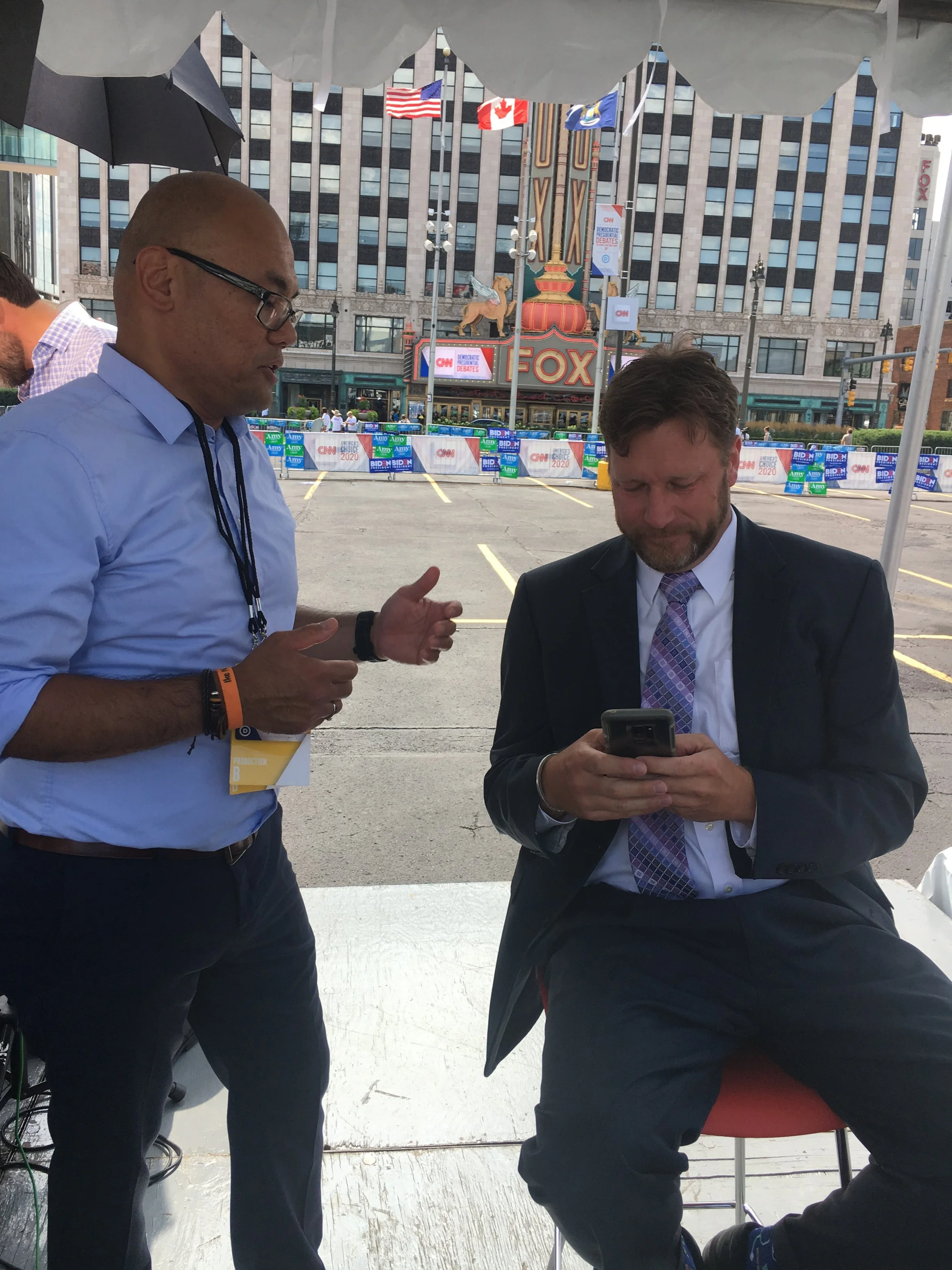 Two men engaged in conversation under a tent in a parking lot, with the Fox Theatre and campaign signs in the background.