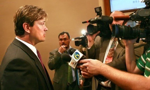 Man in black suit being interviewed by reporters with cameras and microphones