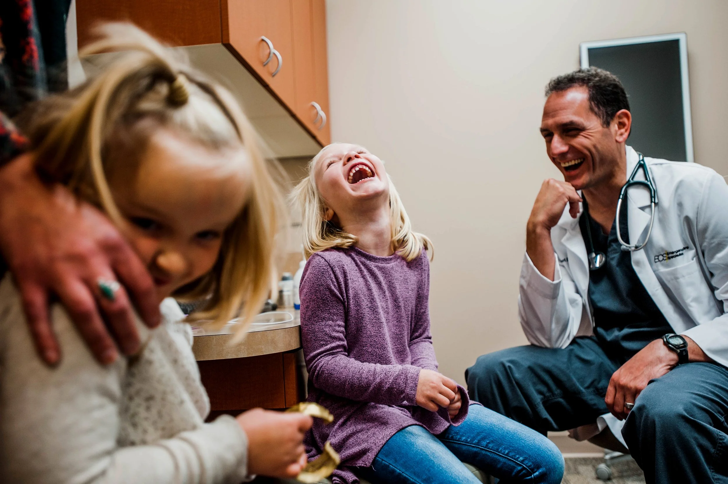 A joyful scene inside a medical office with two young girls, one in the foreground and one being examined, laughing with a doctor in a white coat. The doctor is laughing along with the girls.