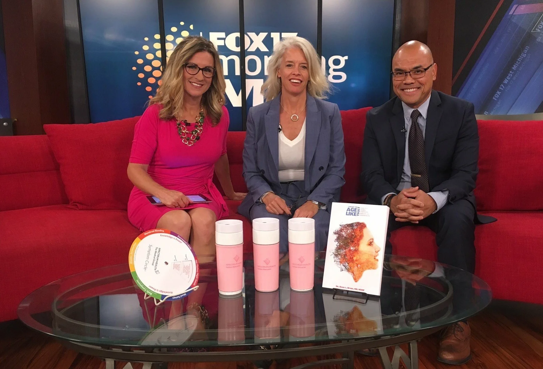 Three news anchors sitting on a red couch in a TV studio with a FOX 17 backdrop. The woman on the left is wearing a pink dress and glasses, holding a tablet. The woman in the middle has platinum blonde hair, is dressed in a blue blazer and white top, and is smiling. The man on the right is bald, wearing glasses, a dark suit, and a patterned tie. In front of them on a glass table are four pink insulated cups