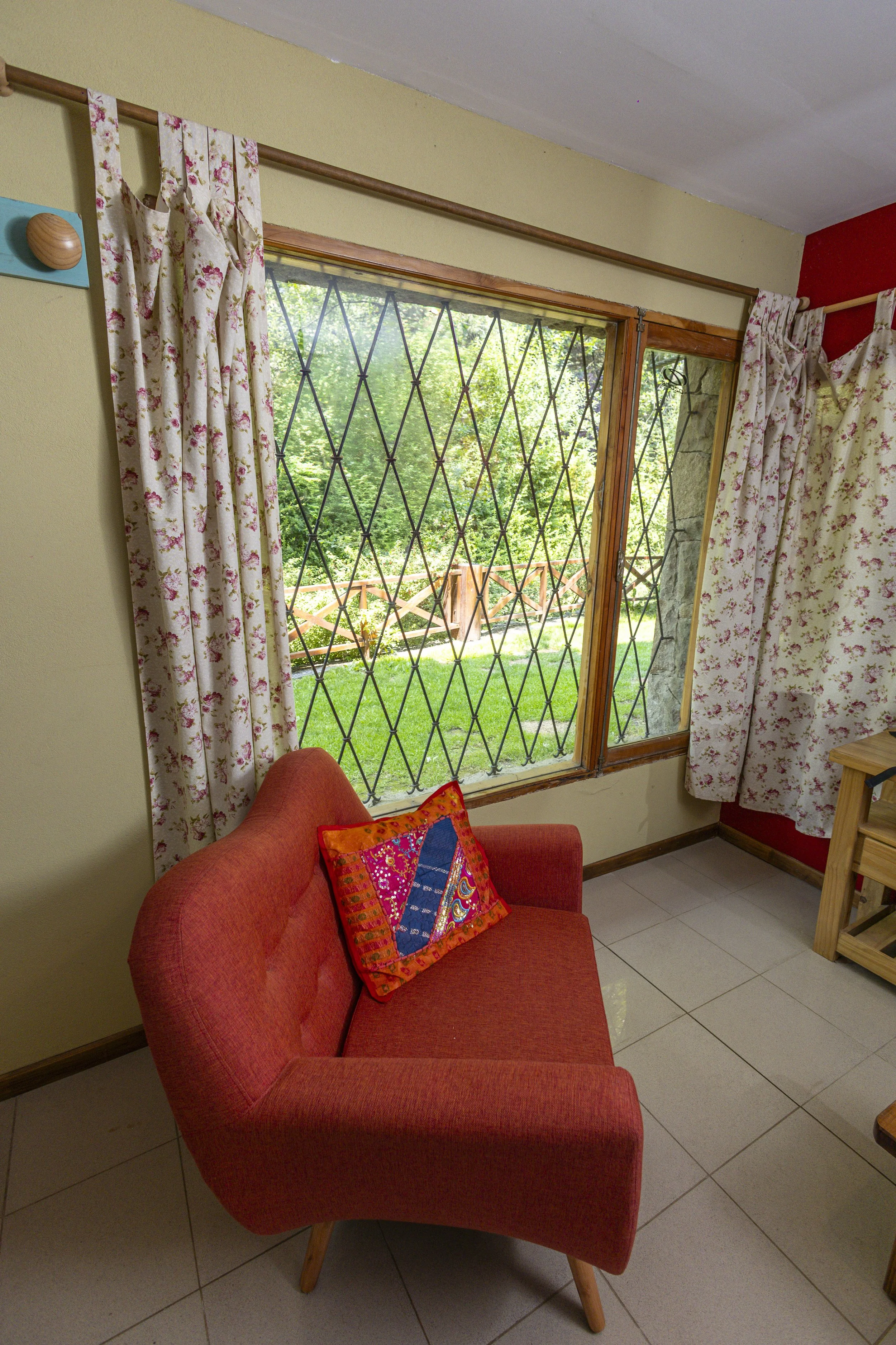 A cozy living room corner with a dusty rose-colored armchair, decorated with a vibrant embroidered pillow. Behind, a large window with diamond-patterned security bars reveals a lush green garden with a wooden fence.