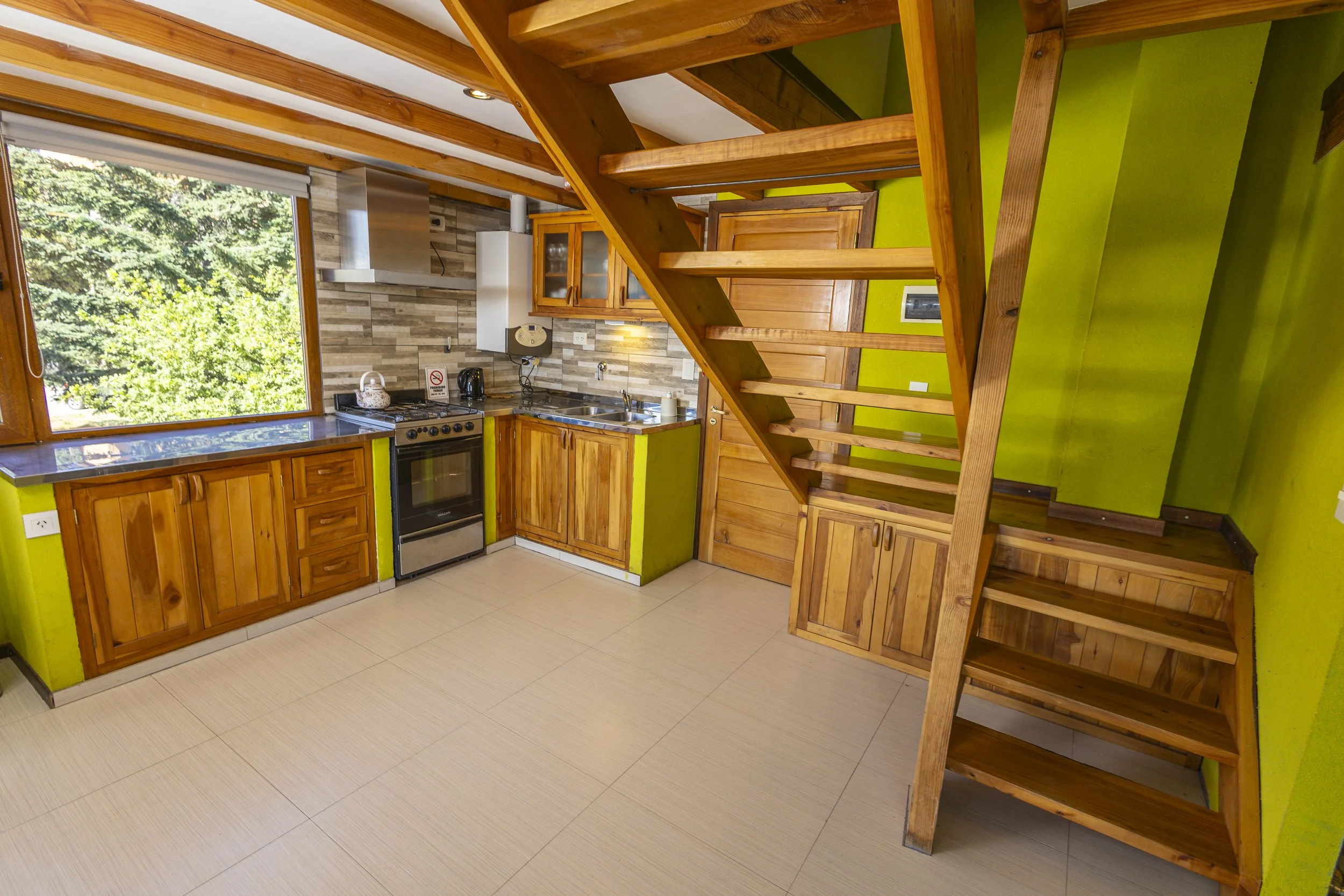 A small kitchen with wooden cabinets, a stove, and a large window overlooking trees, with a wooden staircase next to the wall.