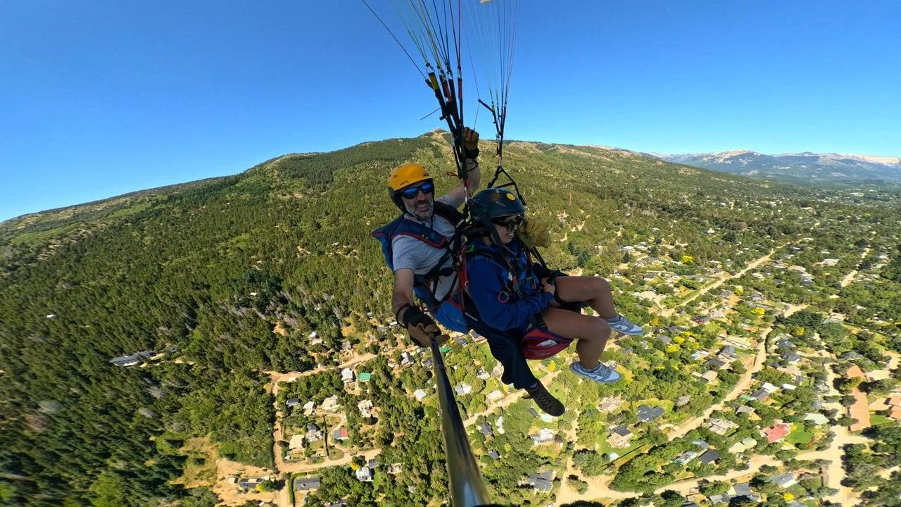 Two people tandem paragliding over a forested town with a mountain background under a clear blue sky.