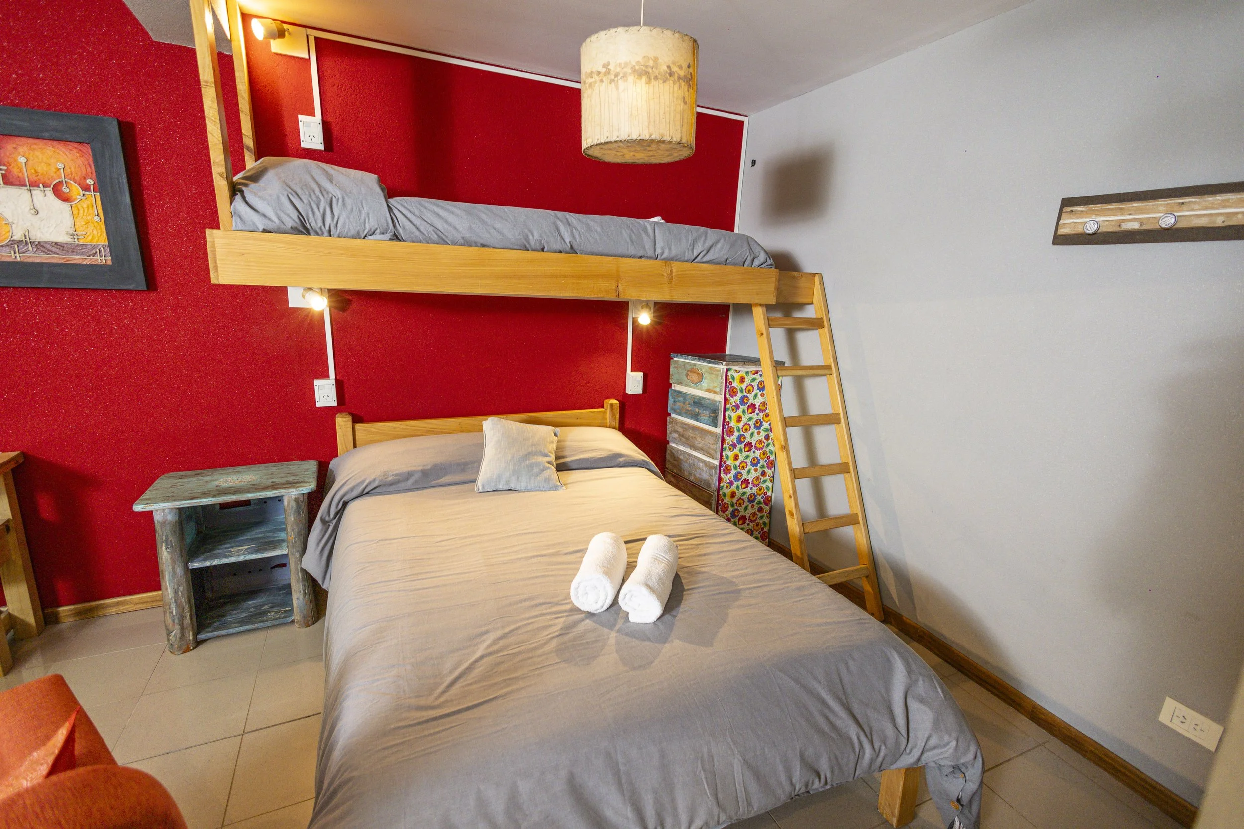 Room with a red accent wall featuring a bed with a wooden frame and gray bedding, a bunk bed with a ladder, a small rustic side table, and a colorful painted drawer, with framed artwork and a hanging light fixture.