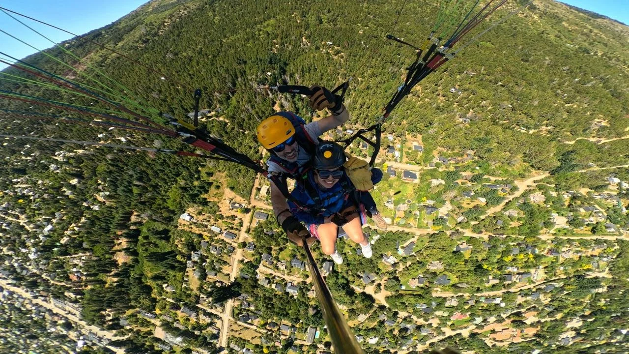 Two people tandem paragliding high above a green, residential area with trees and houses.