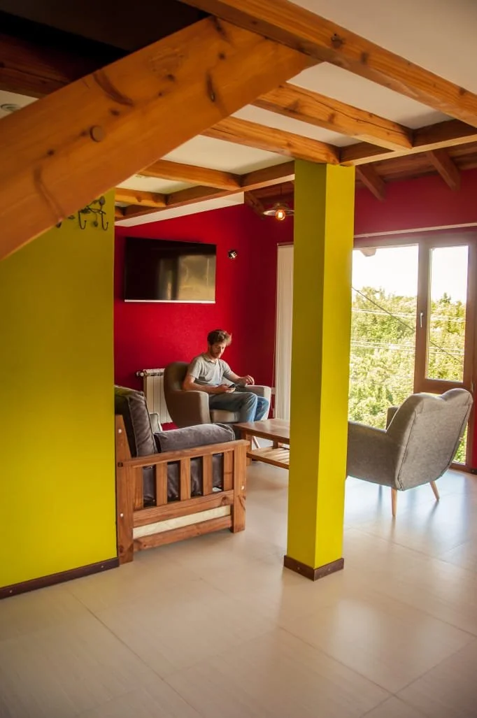 Living room with green and red walls, a person sitting on a brown armchair, wooden ceiling beams, large window with a view of trees, a flat-screen TV on the red wall, and various chairs and furniture.