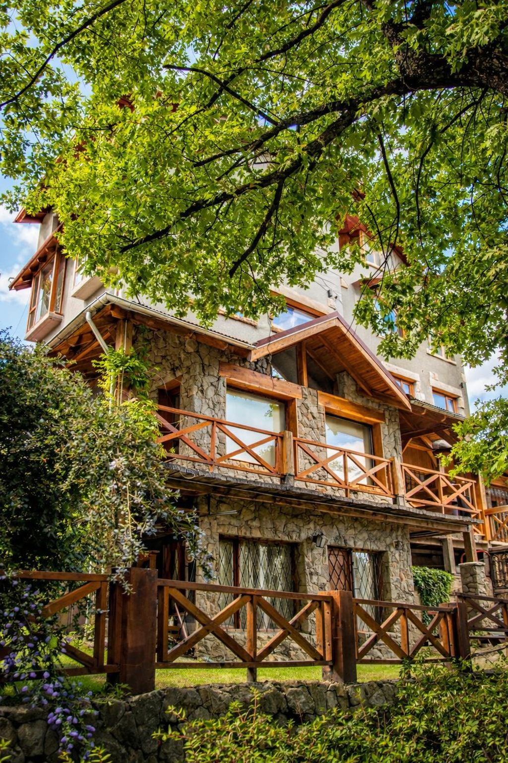 A multi-story house with stone and wood exterior, surrounded by green trees and bushes, with wooden railings on balconies and windows reflecting the sky.