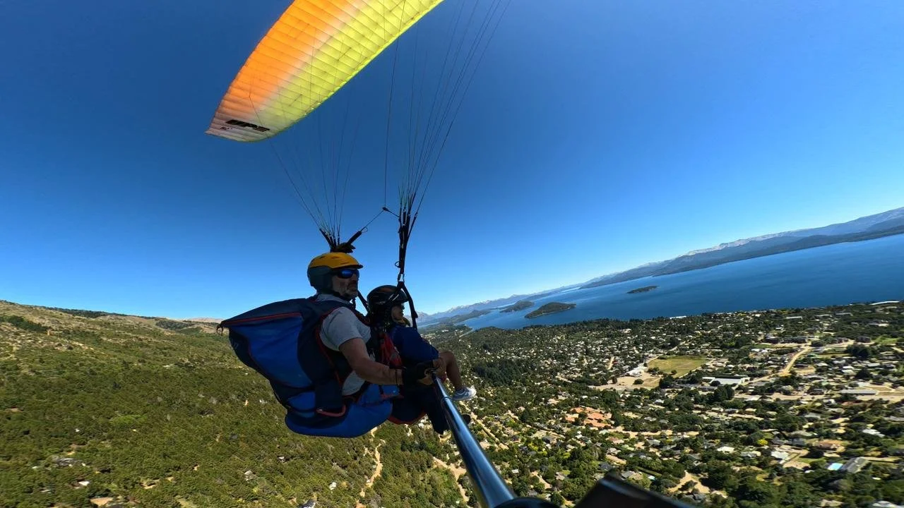 A person and child paragliding over a coastal town with a large body of water and mountains in the background.