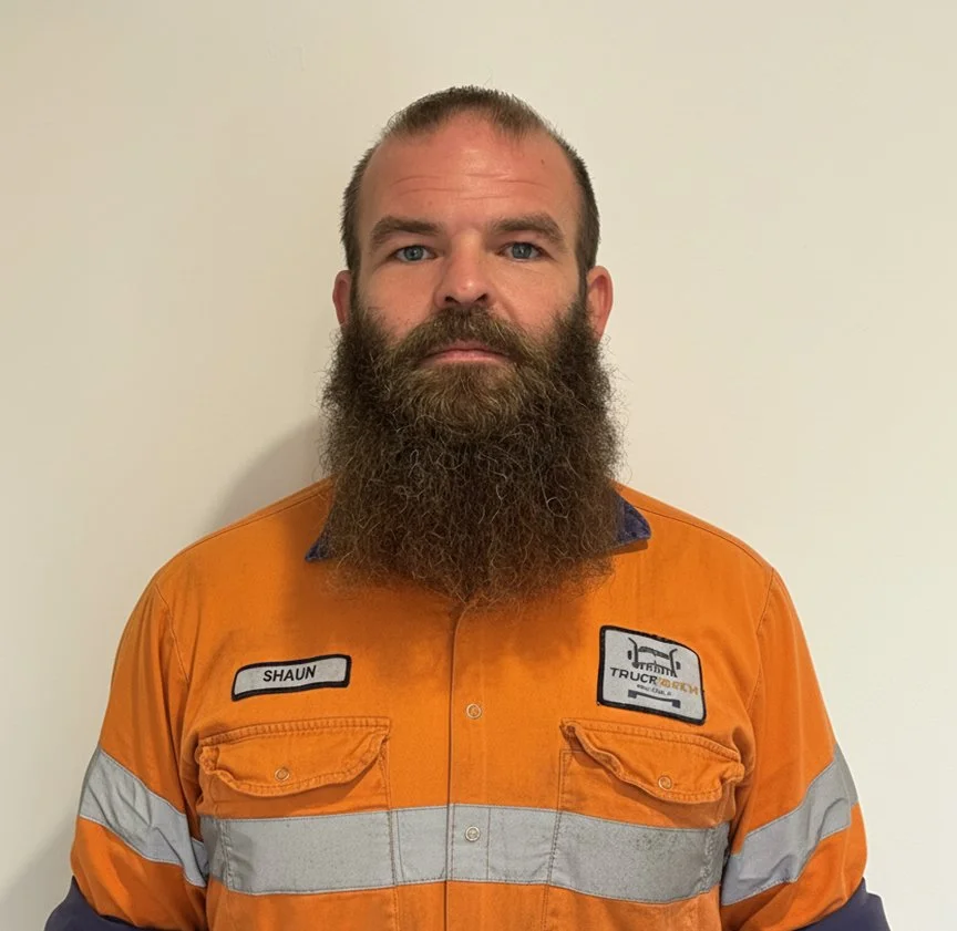 A man with a full beard and short hair wearing an orange safety work shirt with gray reflective stripes, standing against a plain white wall.