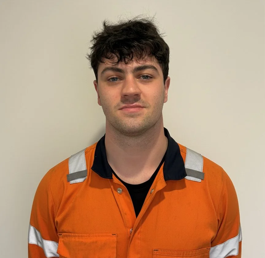 Young man with dark curly hair wearing an orange safety uniform standing against a plain light-colored wall.