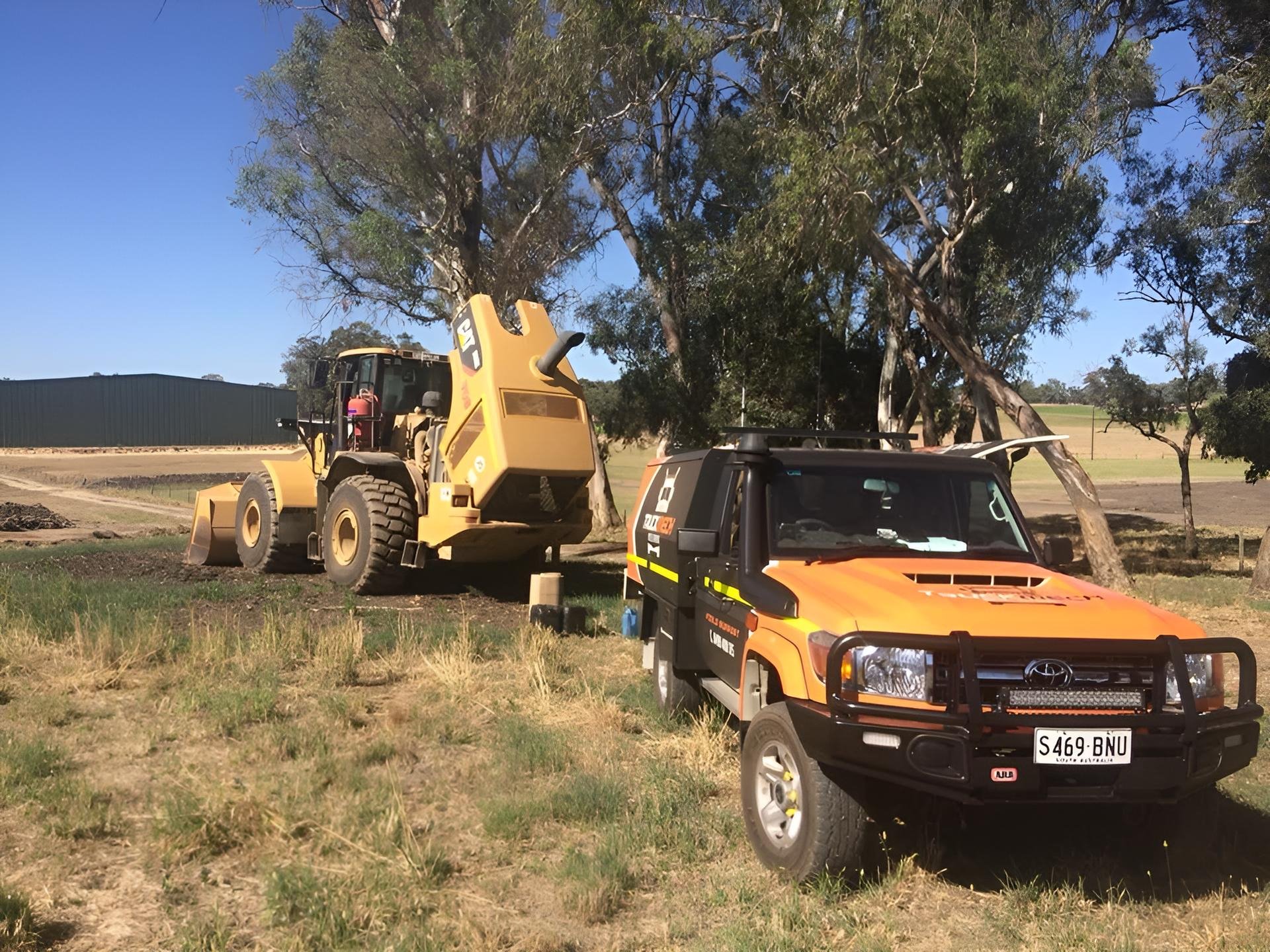 A yellow construction vehicle and an orange off-road vehicle parked on grass near trees and a metal fencing at a construction site.