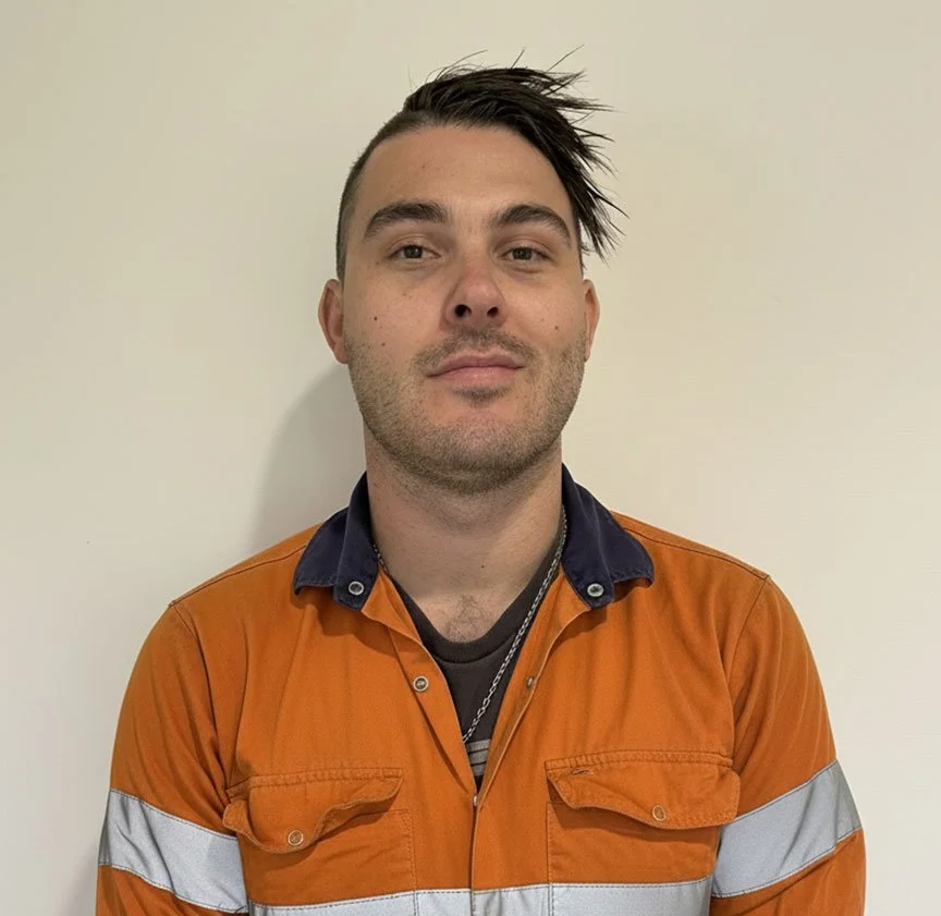 Young man with dark hair styled to one side, wearing an orange safety jacket with reflective stripes, standing against a plain off-white wall.