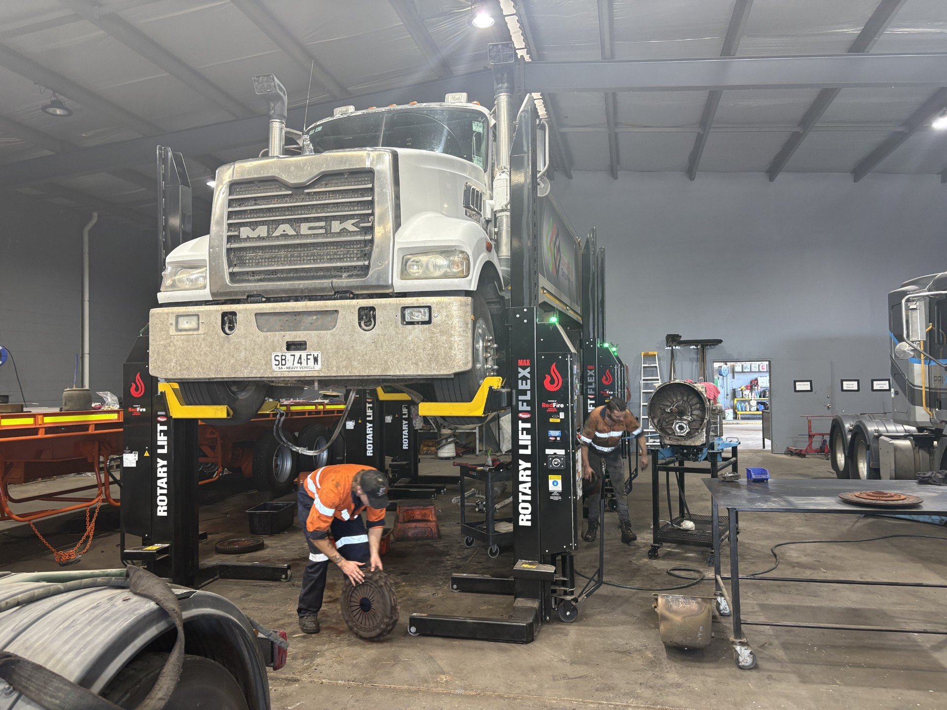 Automotive repair shop with a large Mack truck elevated on a hydraulic lift, mechanics working on the vehicle and various tools and equipment around the workspace.