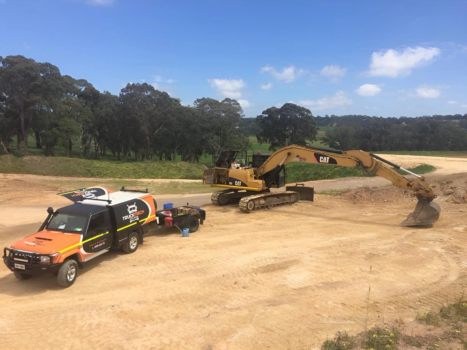 Construction site with excavator and utility truck parked on dirt ground, with green trees and blue sky in the background.