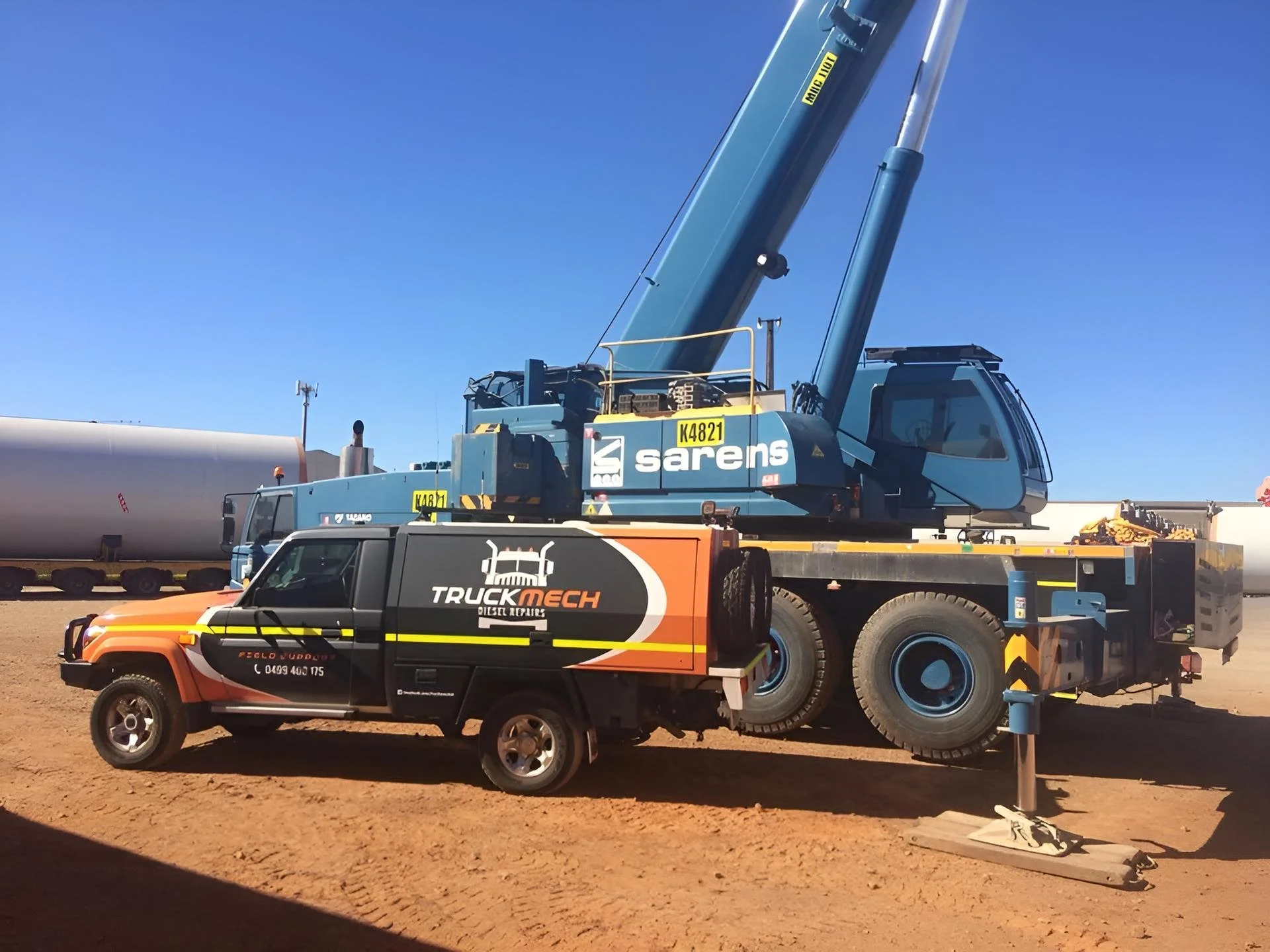Construction site with a large blue crane on a truck lifting a wind turbine blade, and a black pickup truck with