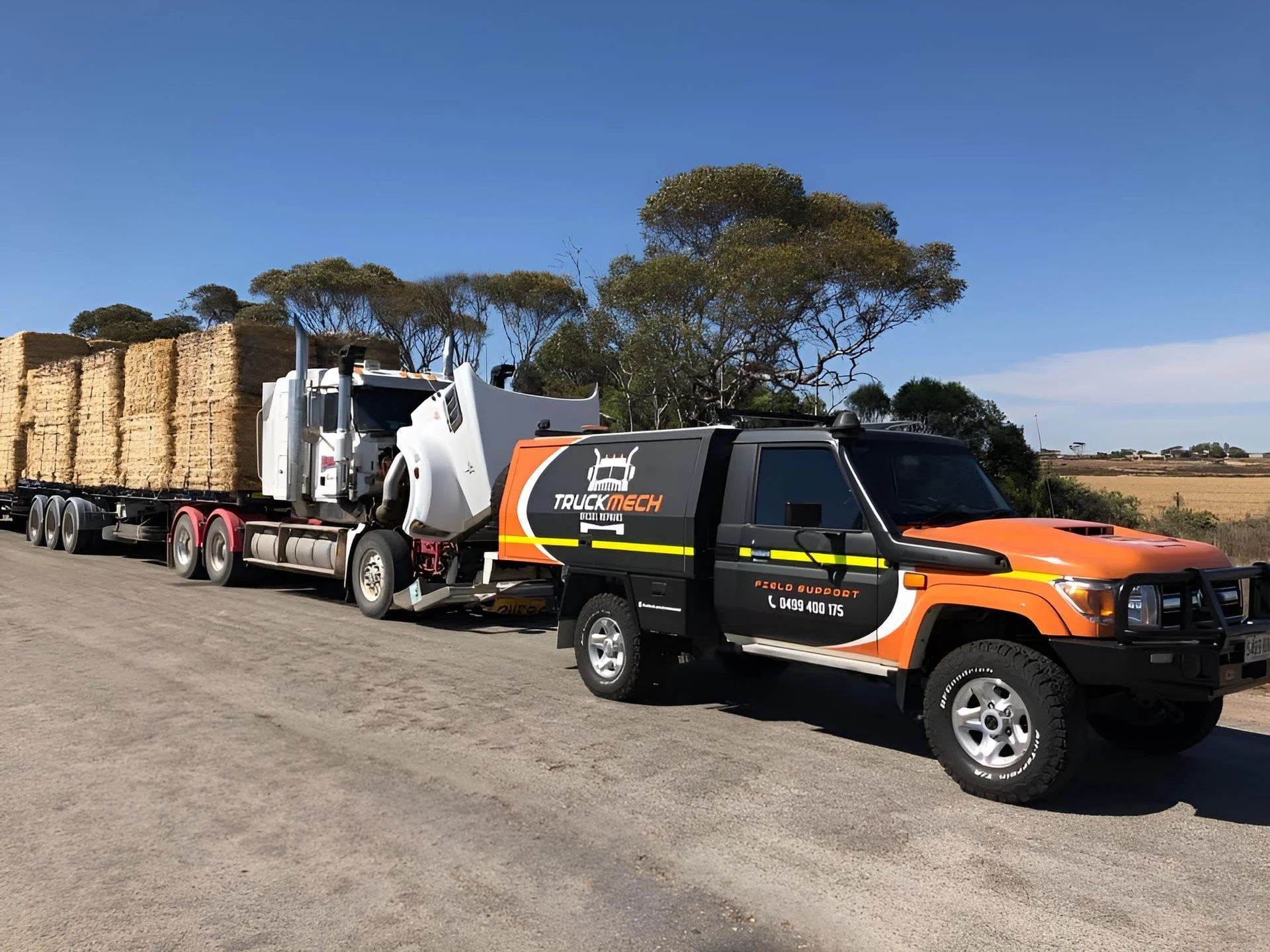 A large orange and black utility truck with a logo reading 'Truck Mech' on the side, pulling a trailer carrying a white semi-truck with a black and orange branding. The semi-truck is carrying a load of hay bales, and the scene is set outdoors with trees and a clear blue sky in the background.
