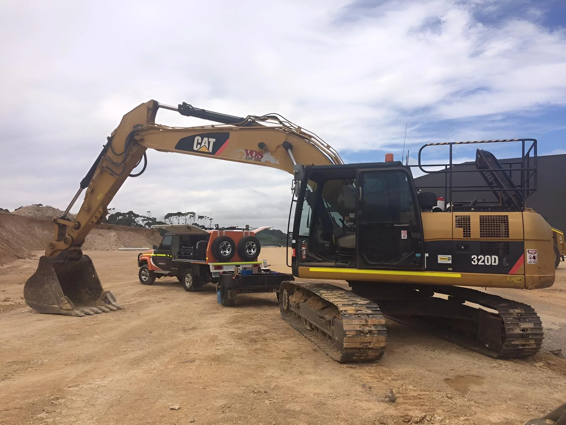 A large yellow Caterpillar excavator with tracks, parked on a dirt construction site, with a small orange and black utility vehicle and a tool box nearby, under a partly cloudy sky.