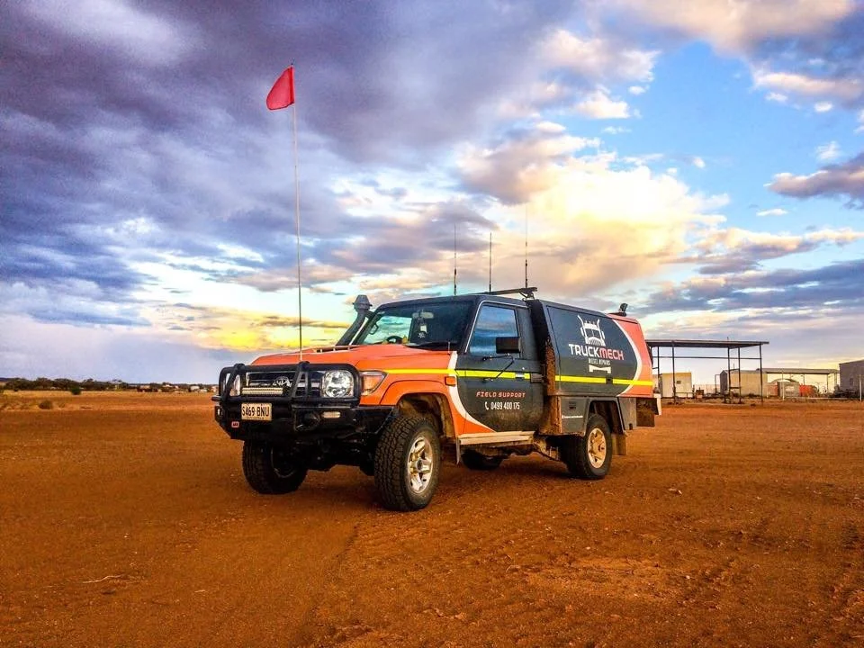 A black and orange utility vehicle with a 'Truck Tech' logo, parked on a reddish dirt field during sunset, with a red flag on a pole and some structures in the background.