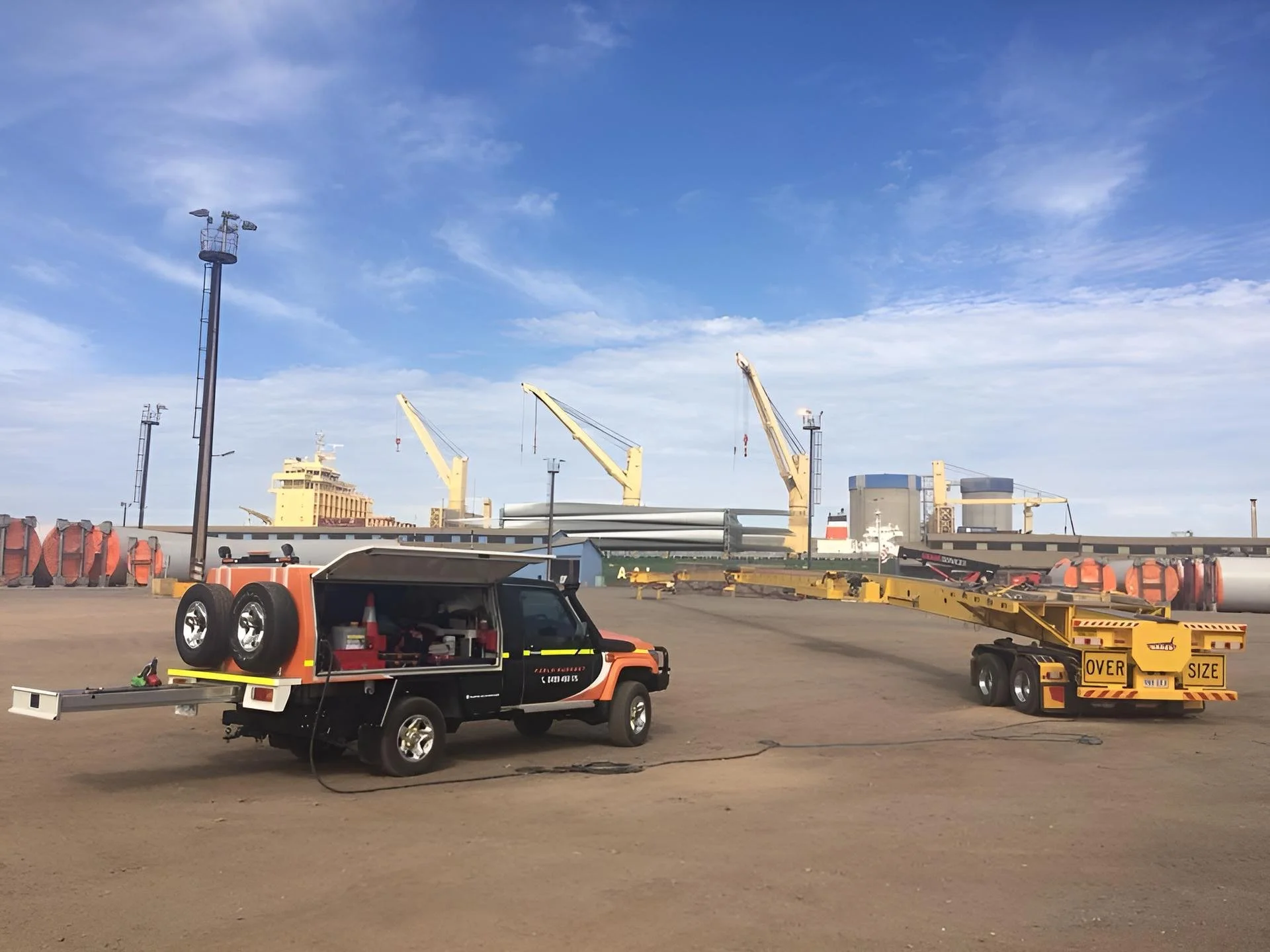 A parking lot with various equipment, including a flatbed truck and a yellow tow truck with a sign that says 'Over Size,' alongside some industrial structures and cranes in the background under a partly cloudy sky.