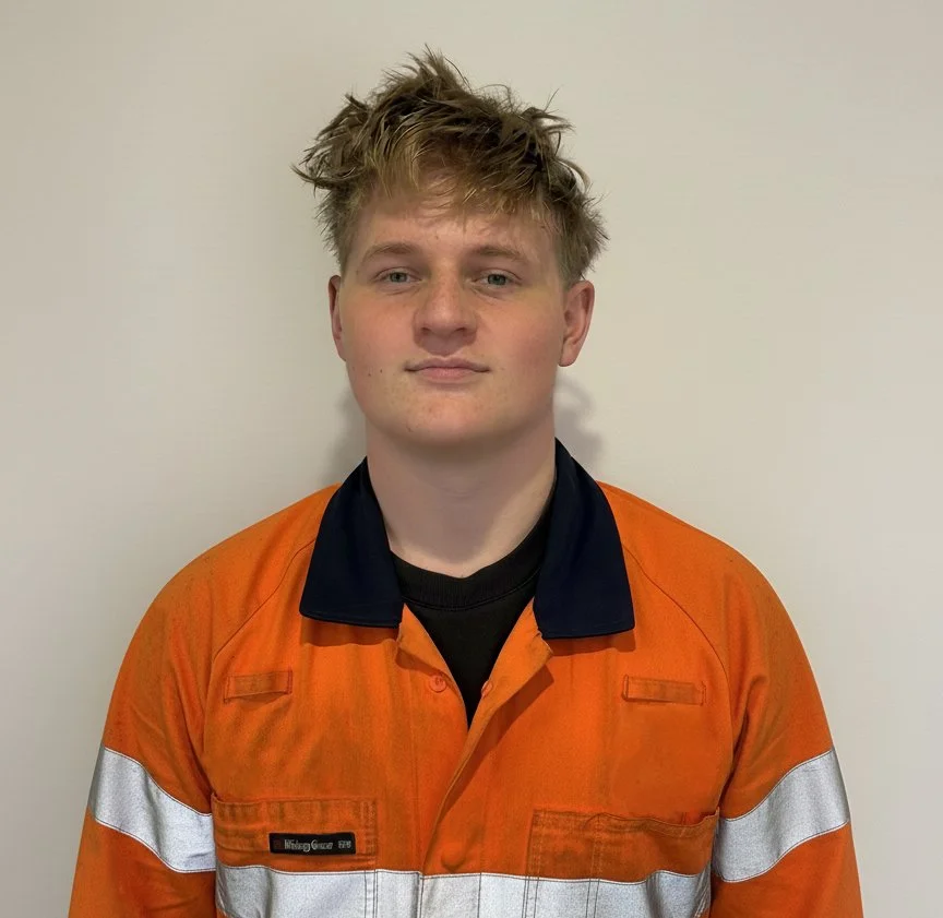 A young man with messy blond hair wearing an orange safety jacket with silver reflective stripes, standing against a plain white wall.