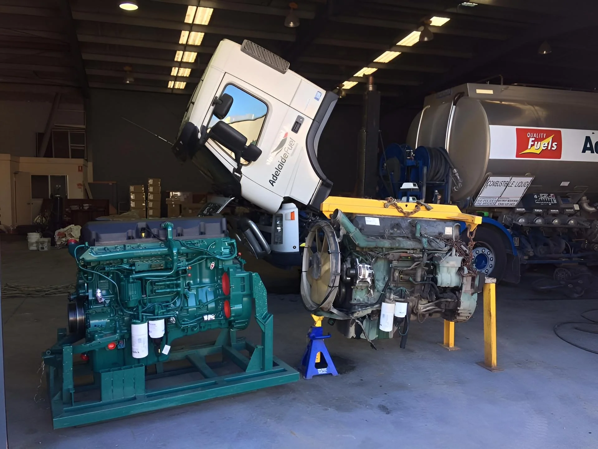 Workshop with a disassembled large green engine, a damaged vehicle cab labeled 'Adelaide Fuel', and a large fuel tank in the background.