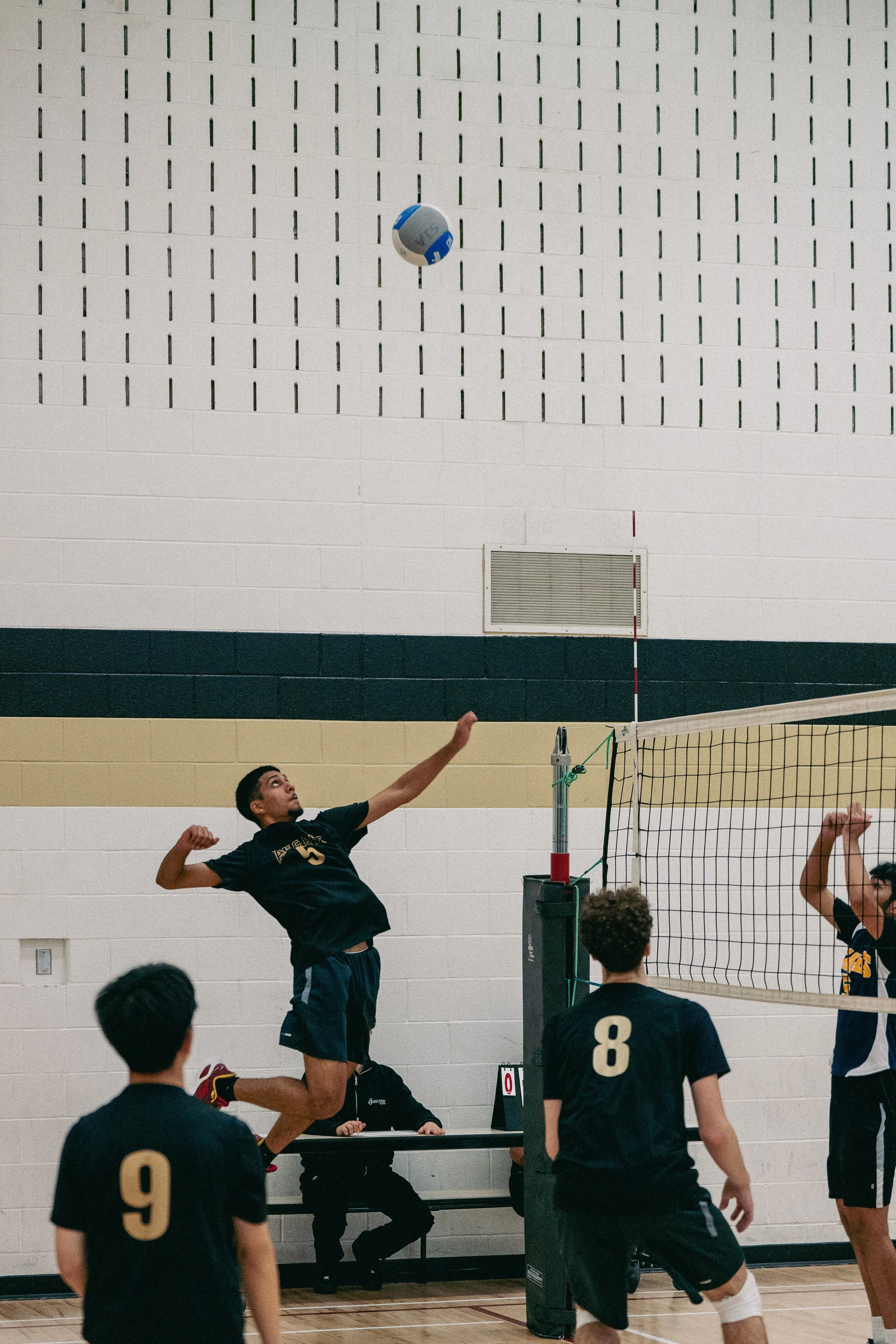 A volleyball player jumps to spike the ball over the net during an indoor game, with teammates ready to defend. The game is played in a gymnasium with a high wall and ventilation vent visible in the background.
