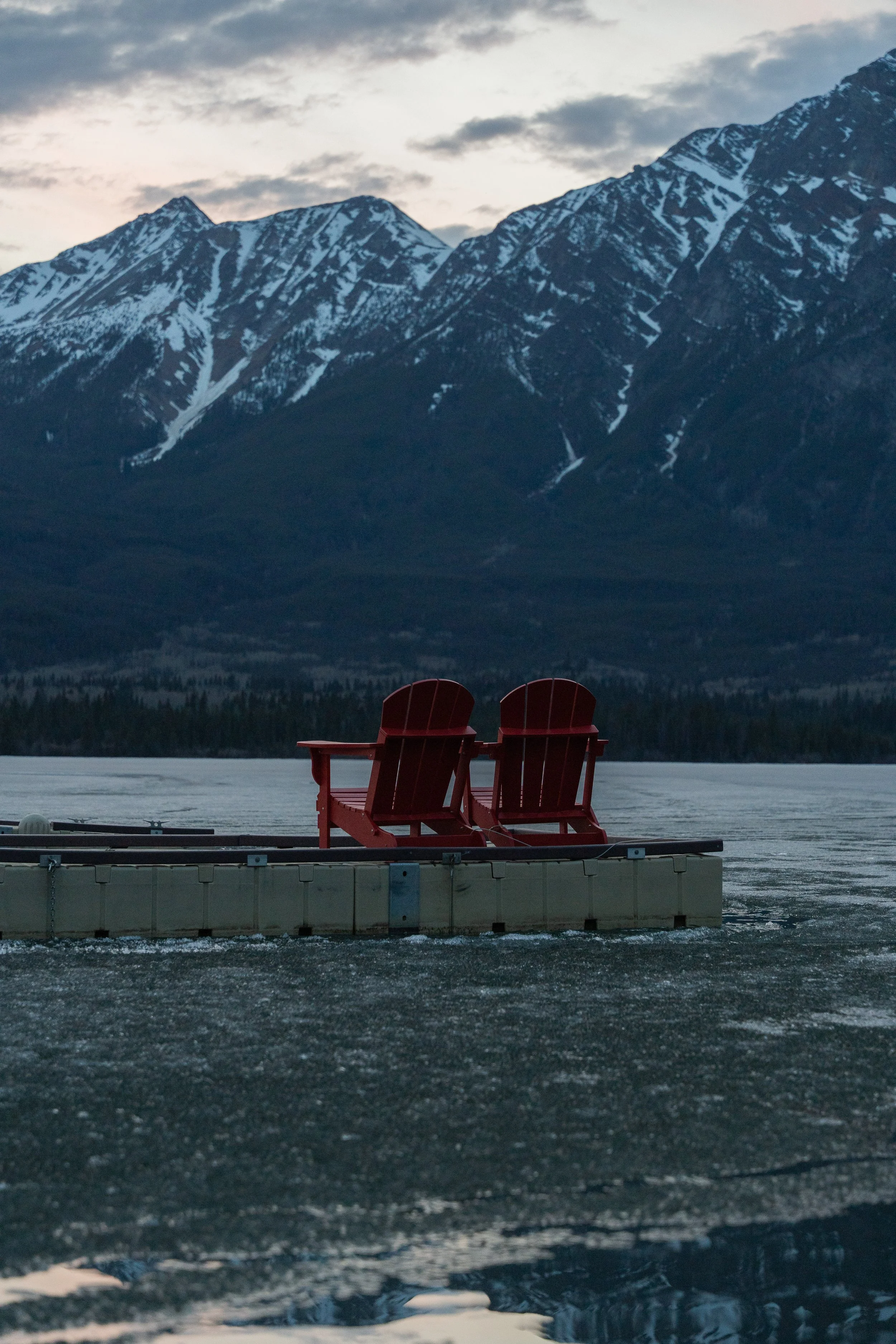 Two red Adirondack chairs on a floating dock on a frozen lake, with snow-capped mountains and an overcast sky in the background.