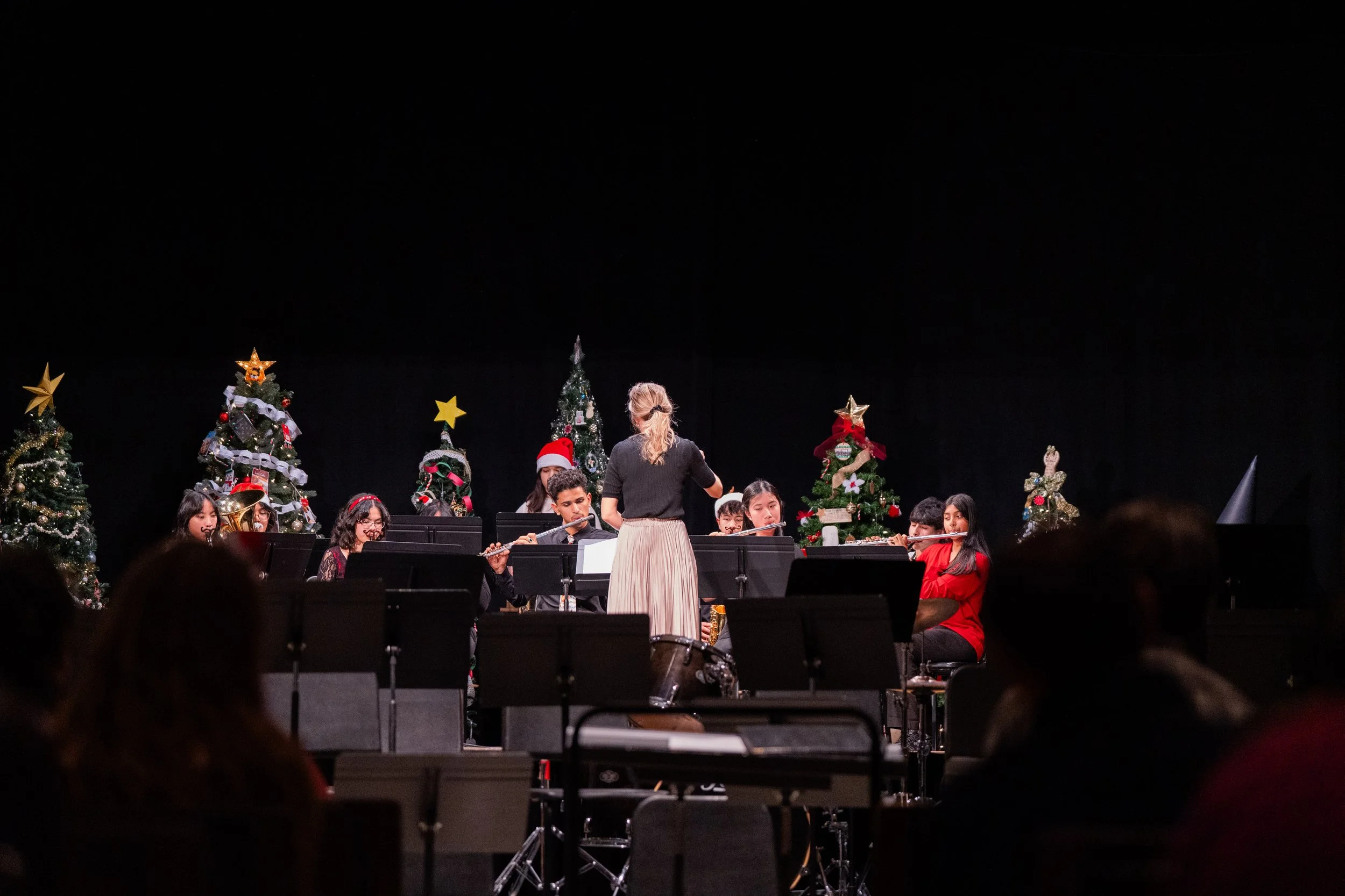 A group of young musicians perform on stage with Christmas trees decorated with ornaments and stars in the background, under a black backdrop, while a conductor leads them.