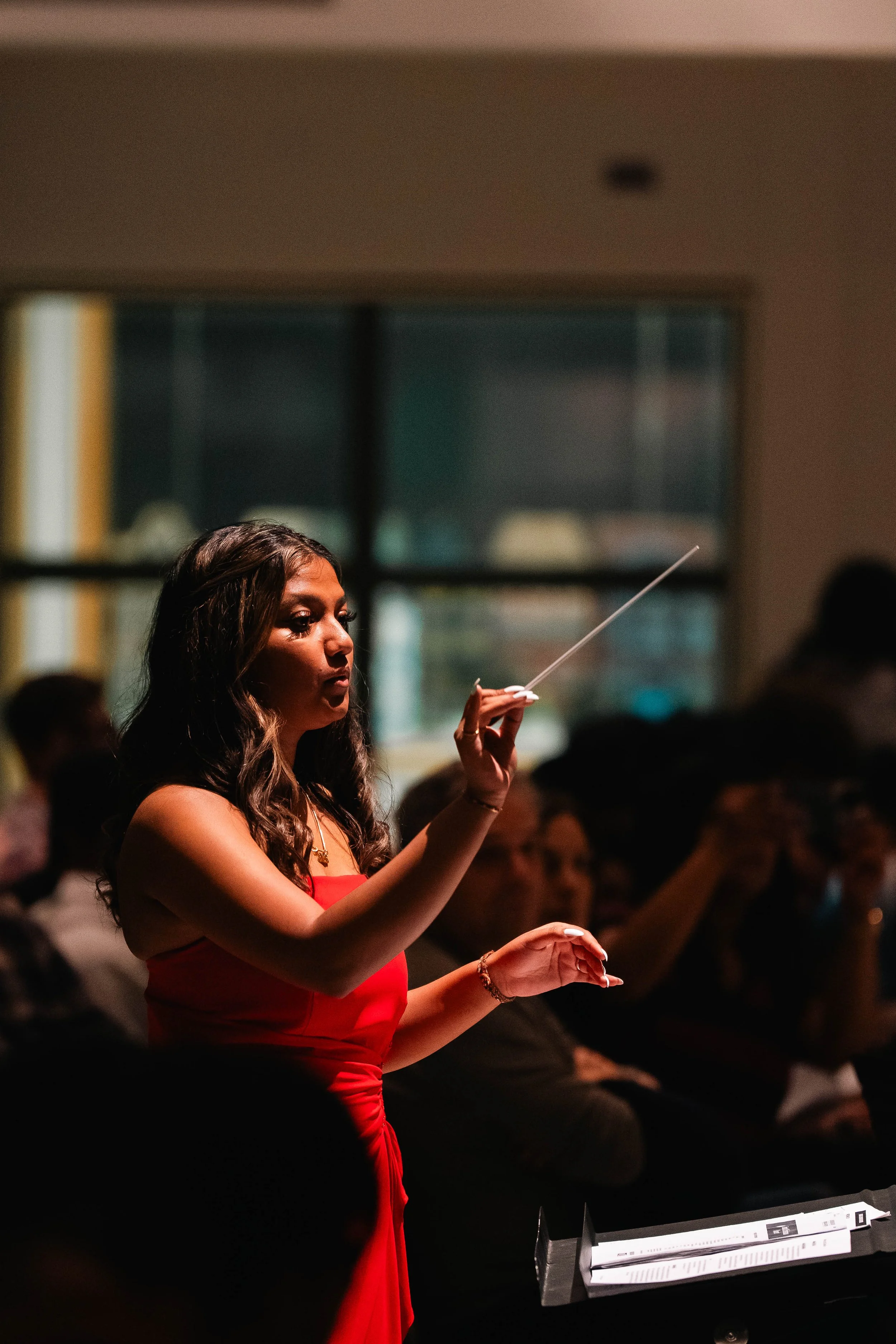 A woman in a red dress conducts an orchestra or choir with a baton in a dimly lit room, with an audience seated in the background.