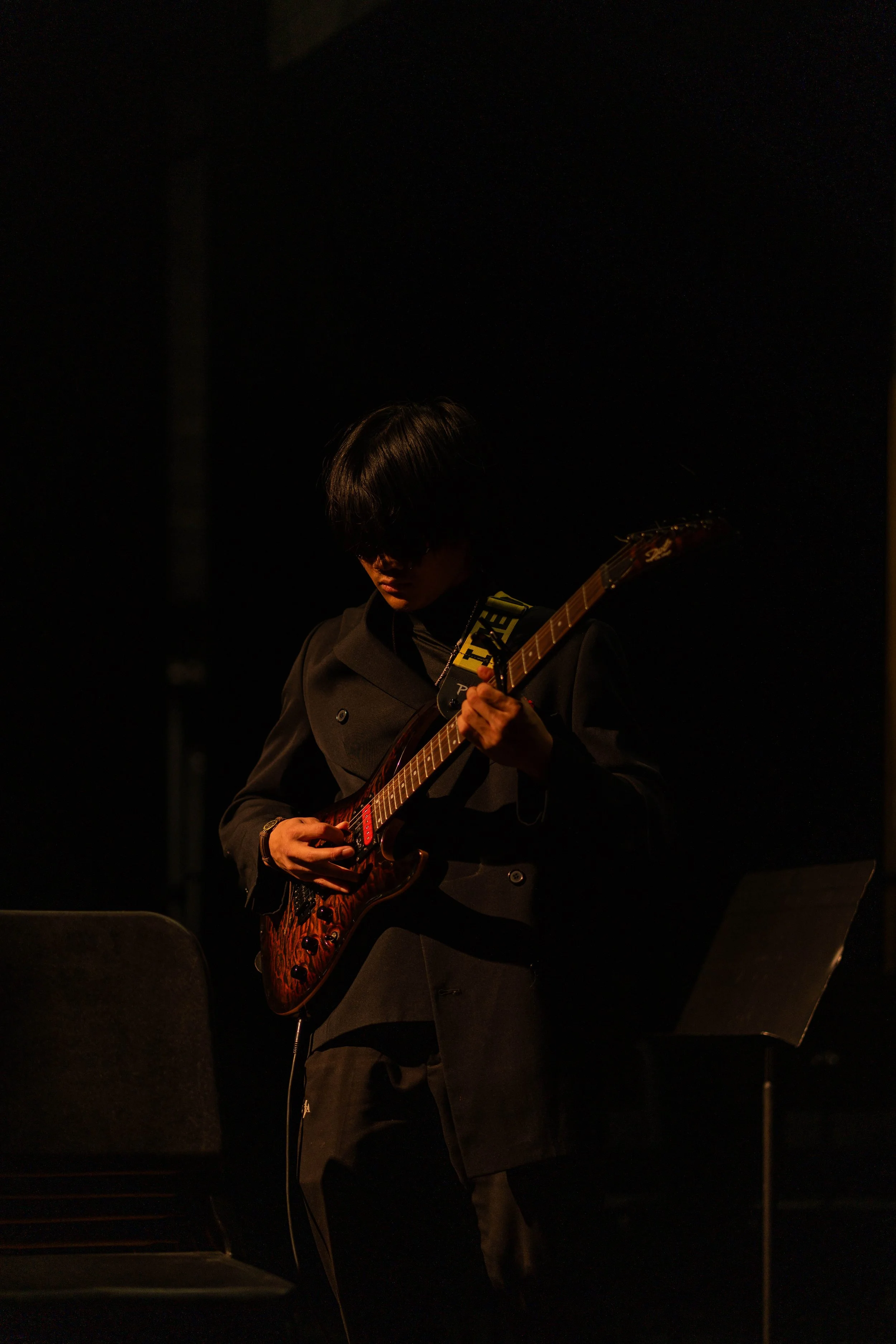 A person playing an electric guitar on stage, dressed in dark clothing, with a music stand nearby and stage lighting creating a moody atmosphere.