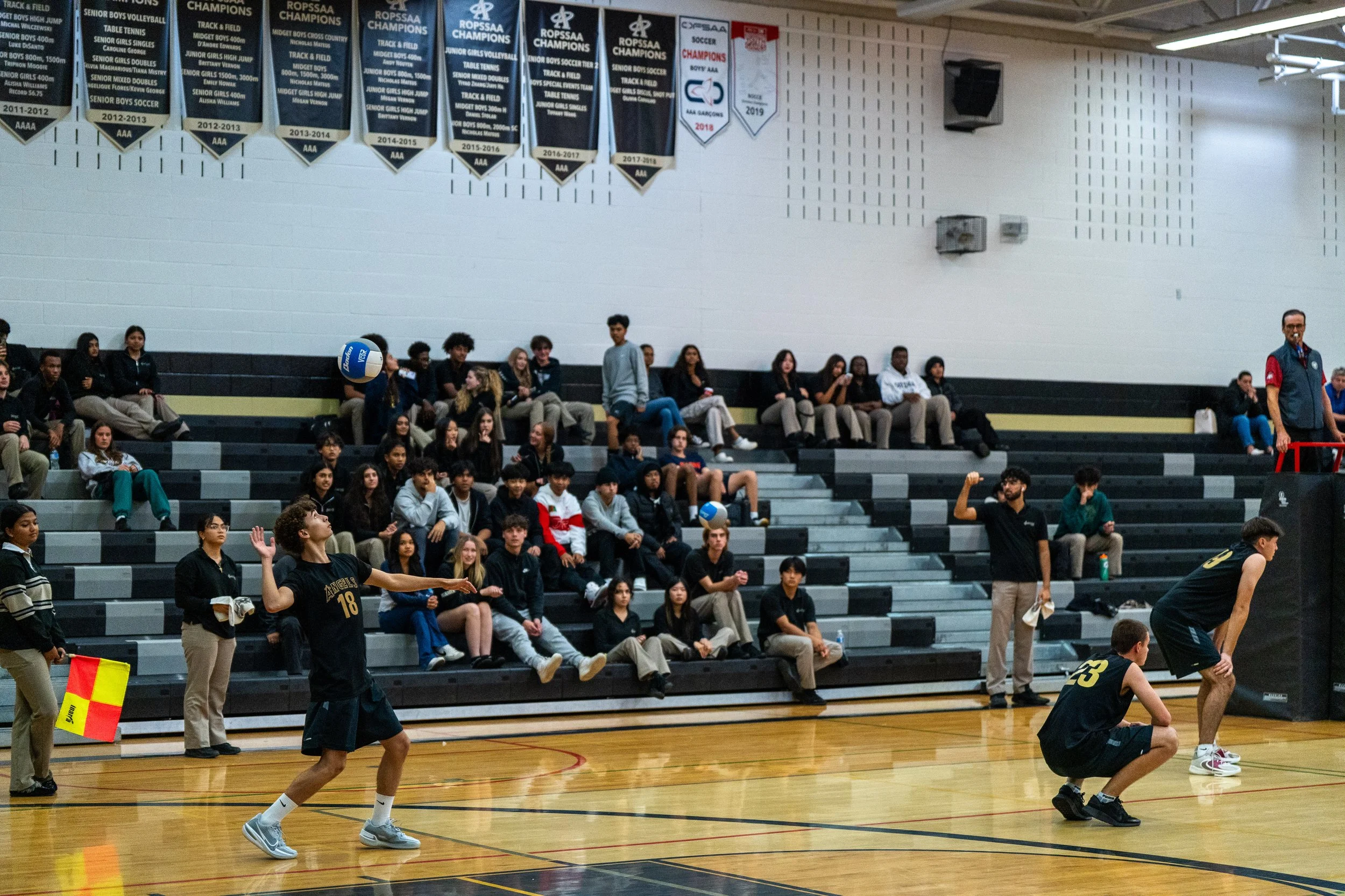 Students playing volleyball in a gymnasium with spectators seated in bleachers and banners hanging on the wall.