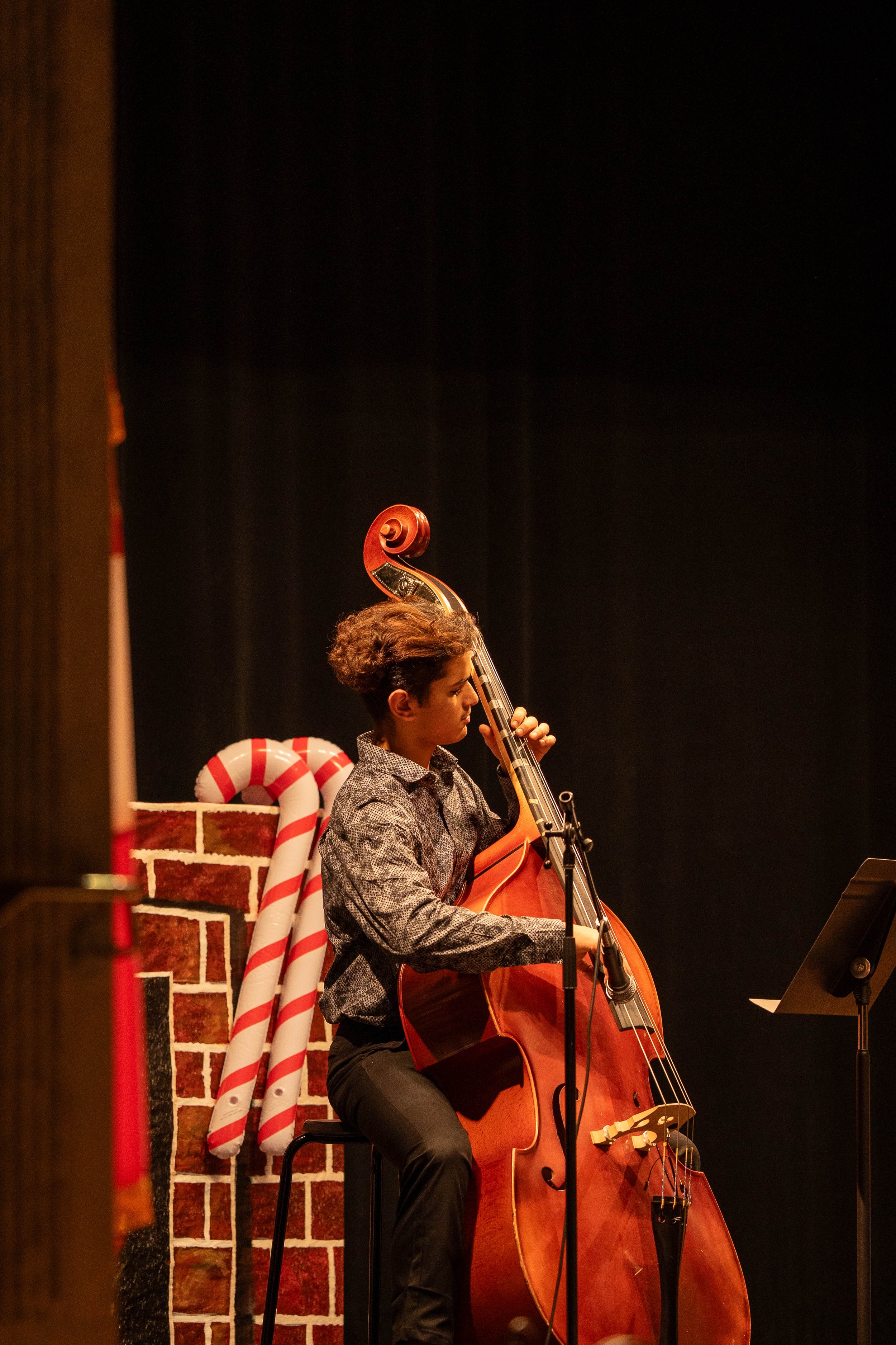 A young person with curly hair, wearing a patterned shirt and black pants, playing a double bass on stage, with a festive holiday Christmas decoration featuring candy canes and a brick fireplace backdrop.