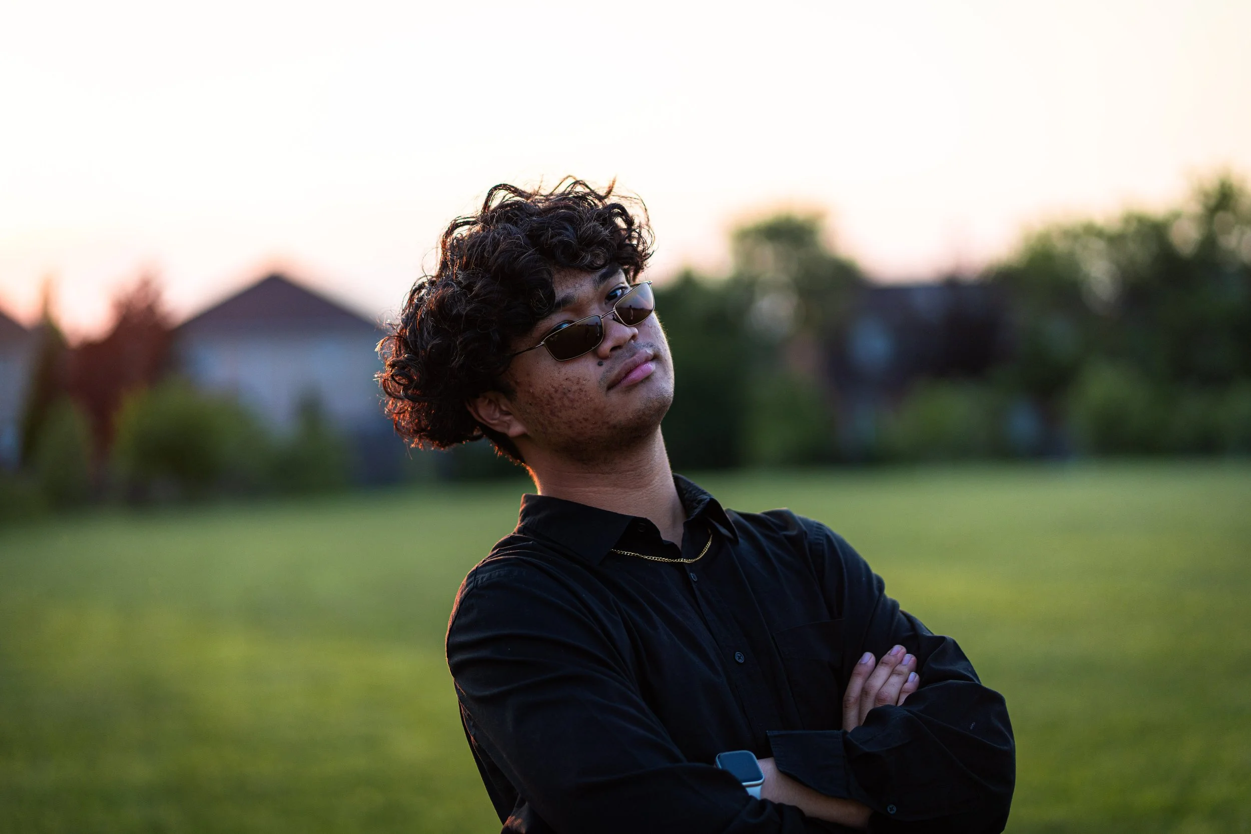 A young man with curly hair, sunglasses, and a black shirt standing outdoors with arms crossed, in a grassy area during sunset.
