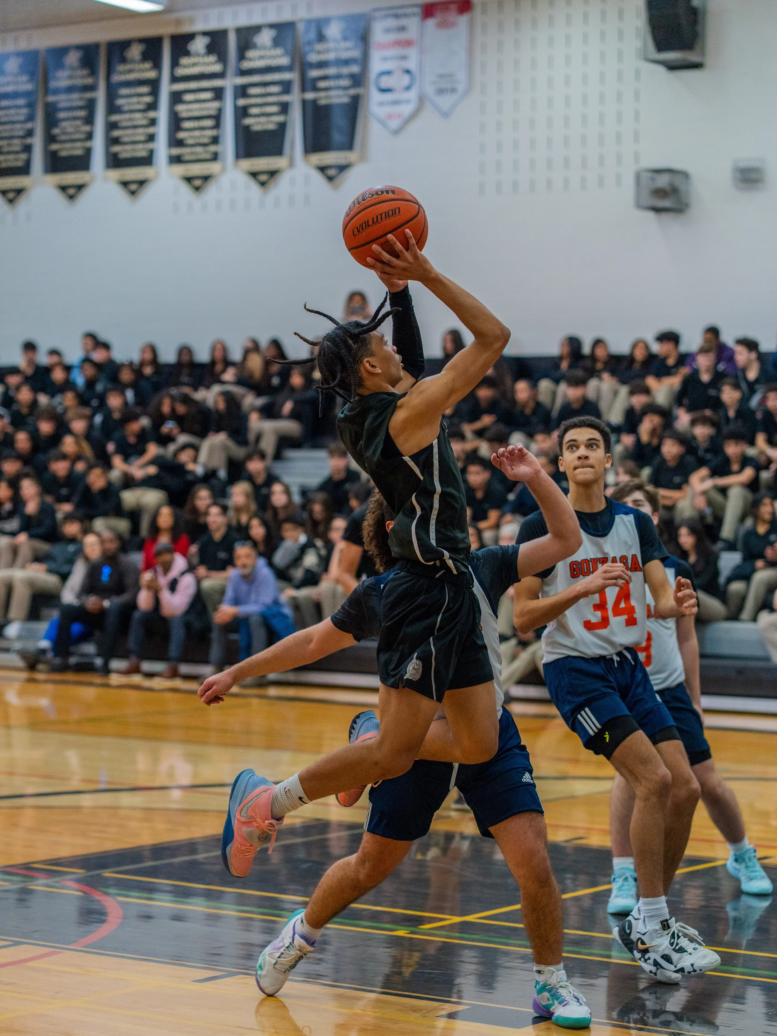 A basketball game with players in action on the court, one player jumping to shoot or pass the ball while others defend, with an audience in the background.