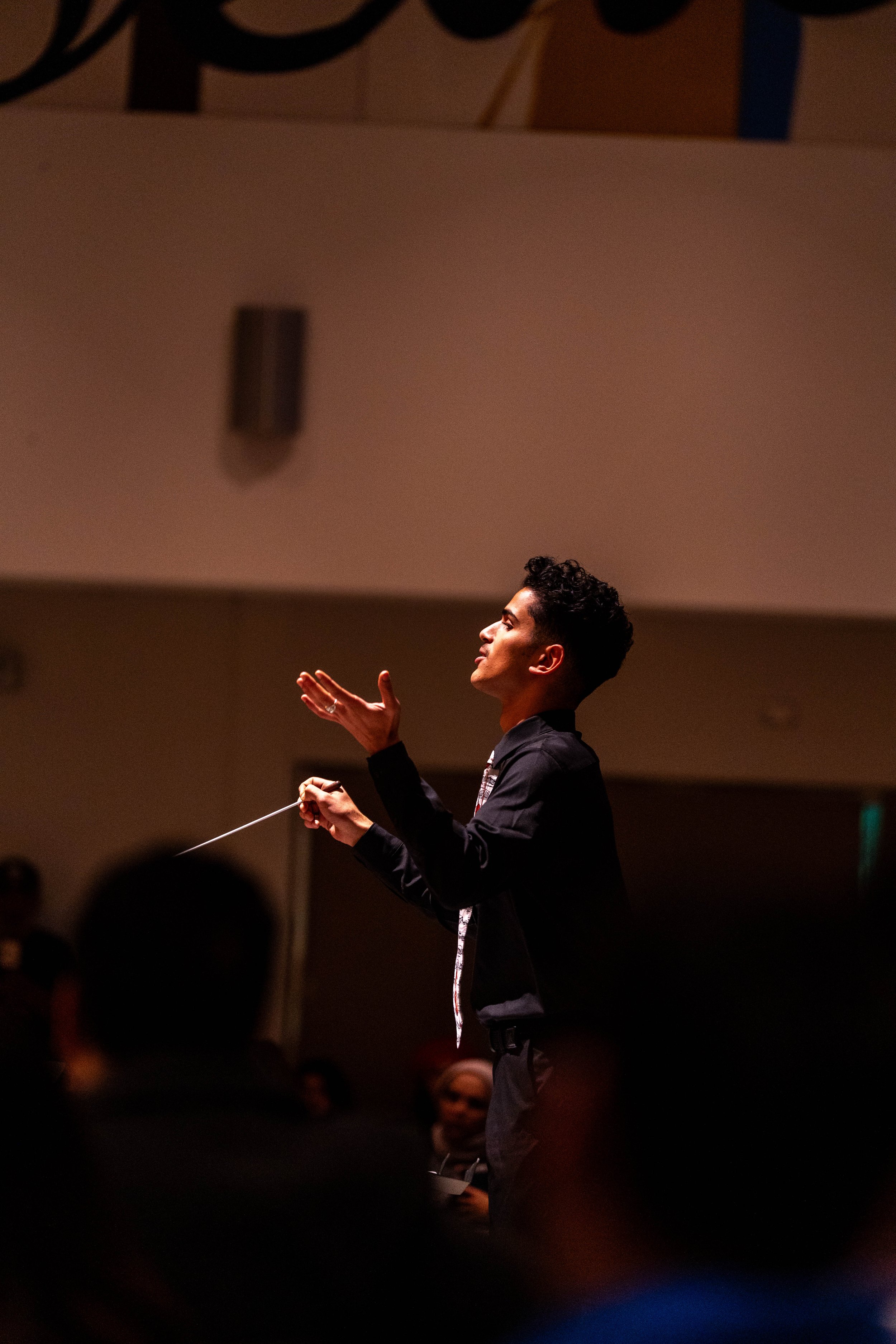 A young man conducting, standing with one hand raised and one hand holding a baton, in a dimly lit concert hall.