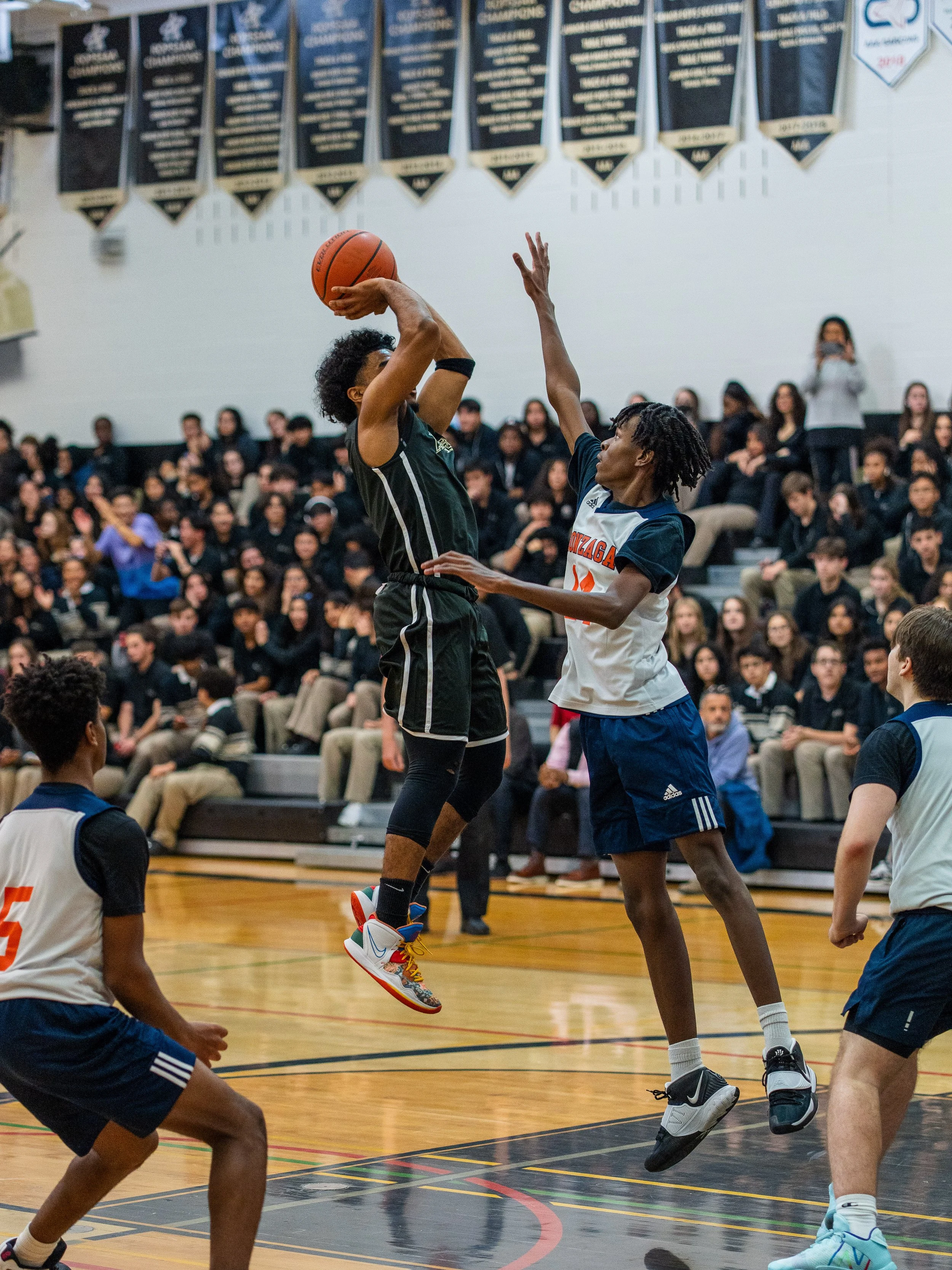 Two young basketball players in action, one jumping to score while the other attempts to block, with a crowd of spectators in the background.