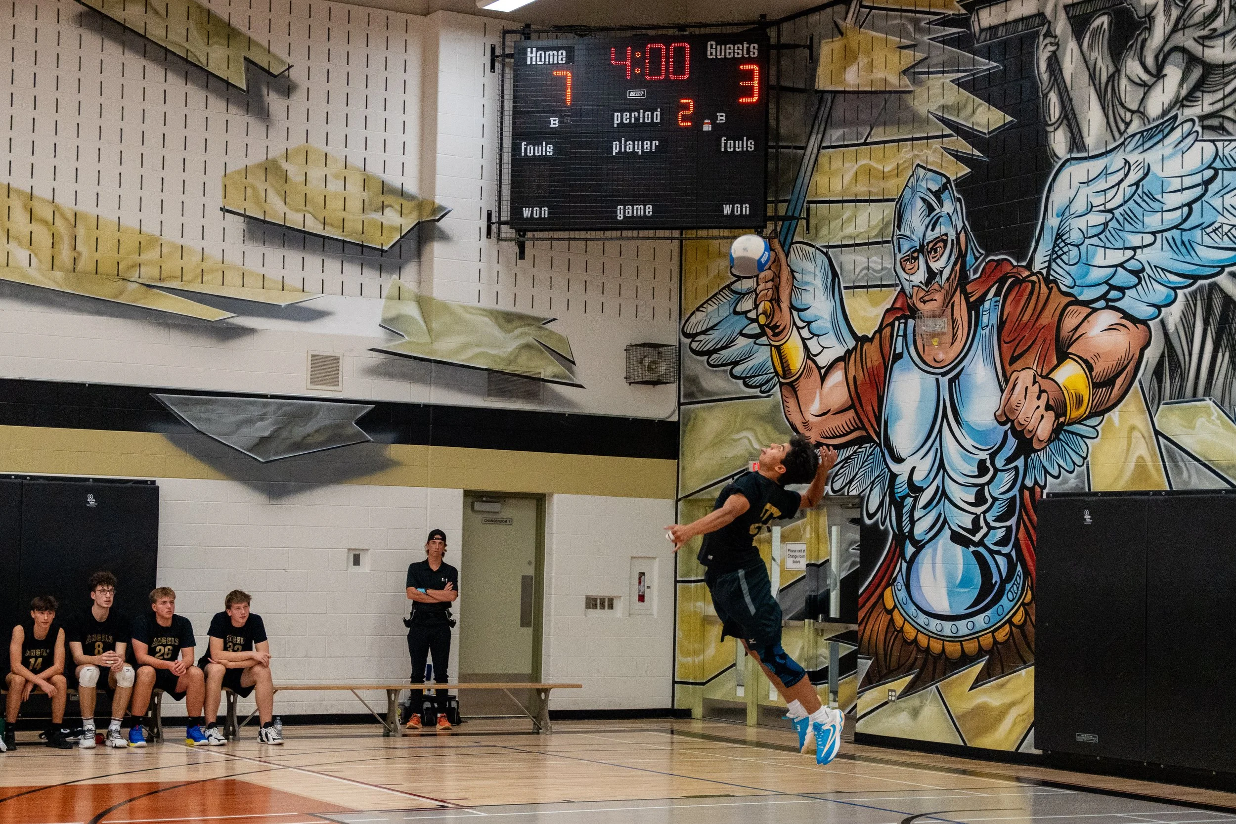 A volleyball player in mid-air spiking the ball at a gymnasium with a colorful wall mural featuring a winged warrior. A scoreboard shows a score of 7-3 with 4 minutes remaining.