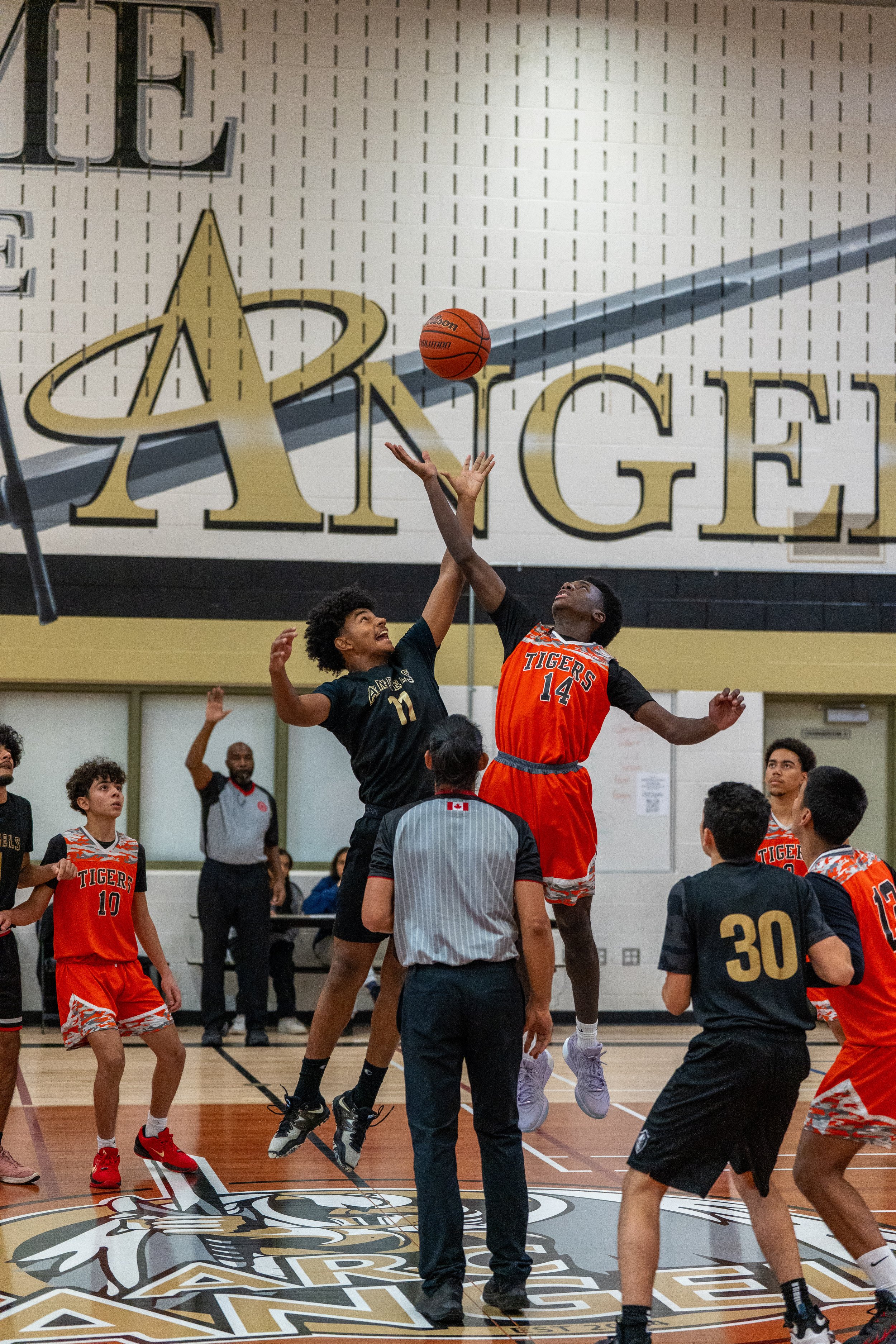 A basketball game in progress with players jumping for the ball at the start of the game, in a gymnasium with a large Lakers logo on the floor and a wall mural reading "ANGE" in the background. Players are in black and orange jerseys, and officials are present on the court.