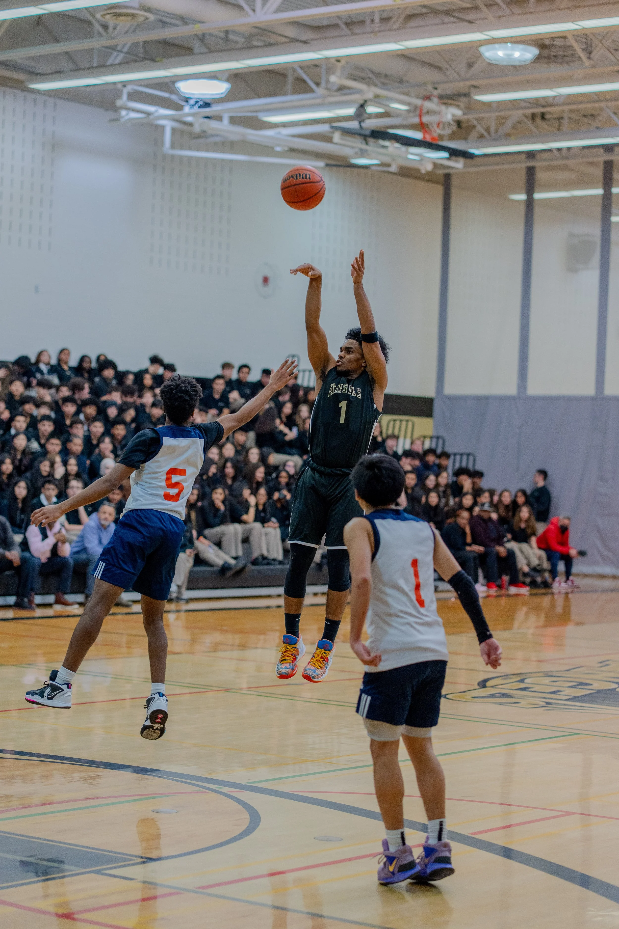 A basketball game in progress with a player in a black uniform jumping to make a shot while an opposing player in a white and blue uniform attempts to block, and another player in a white and blue uniform nearby. The gym is filled with spectators seated on bleachers.