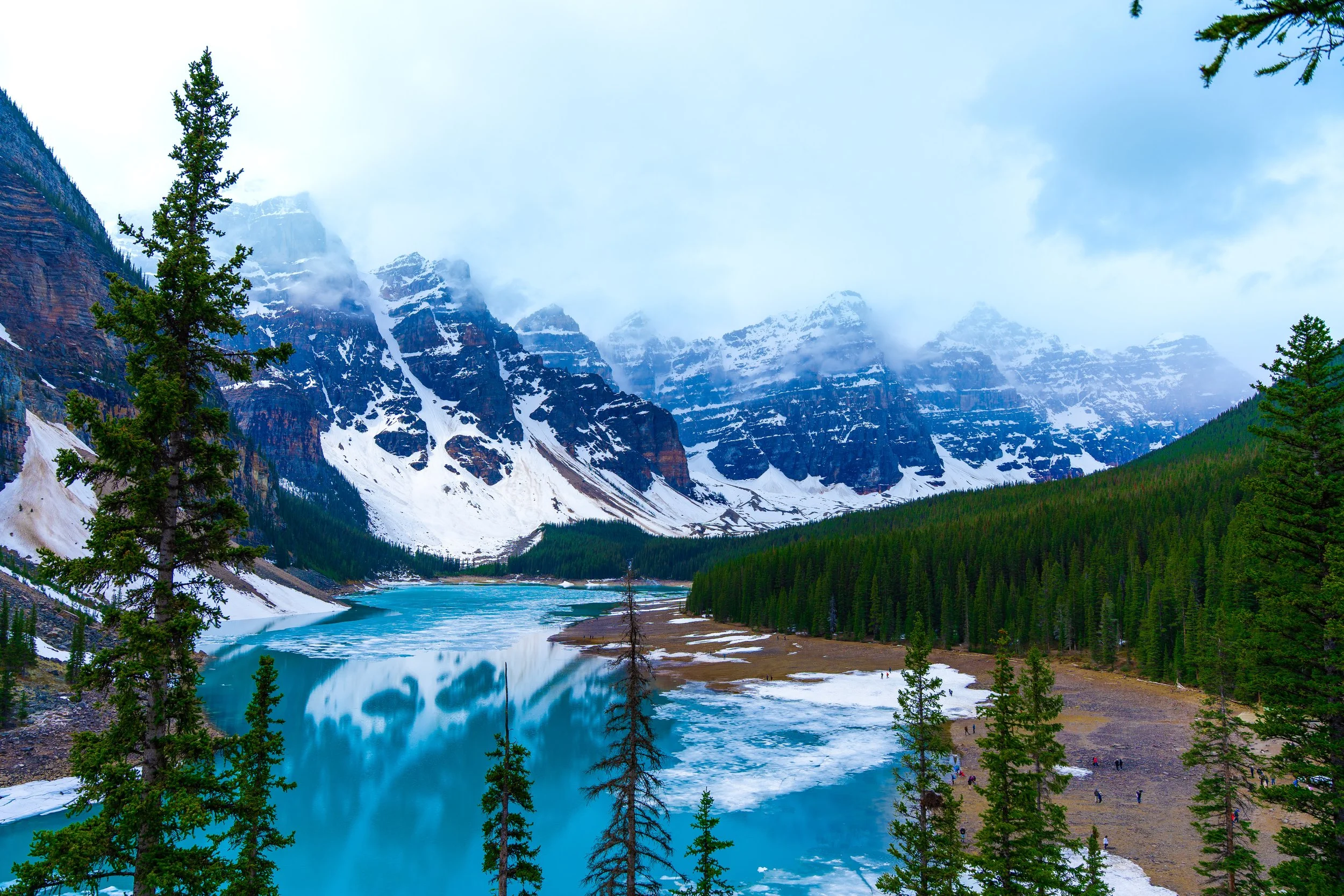 Mountains with snow and clouds, a partly frozen lake, and dense evergreen trees in a serene wilderness