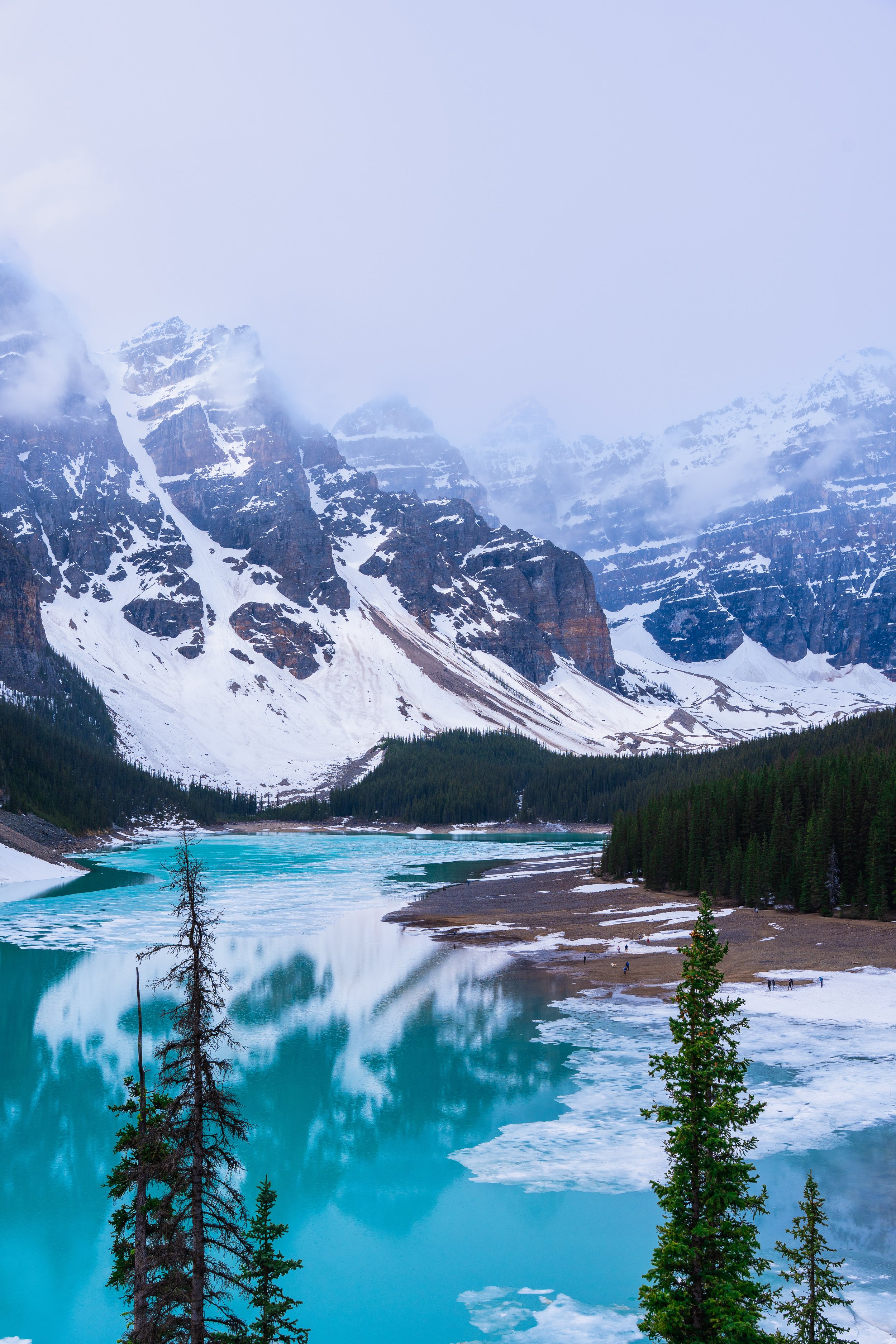Snow-capped mountains surround a turquoise lake with icy patches, dense evergreen forest on the shoreline, and a cloudy sky.