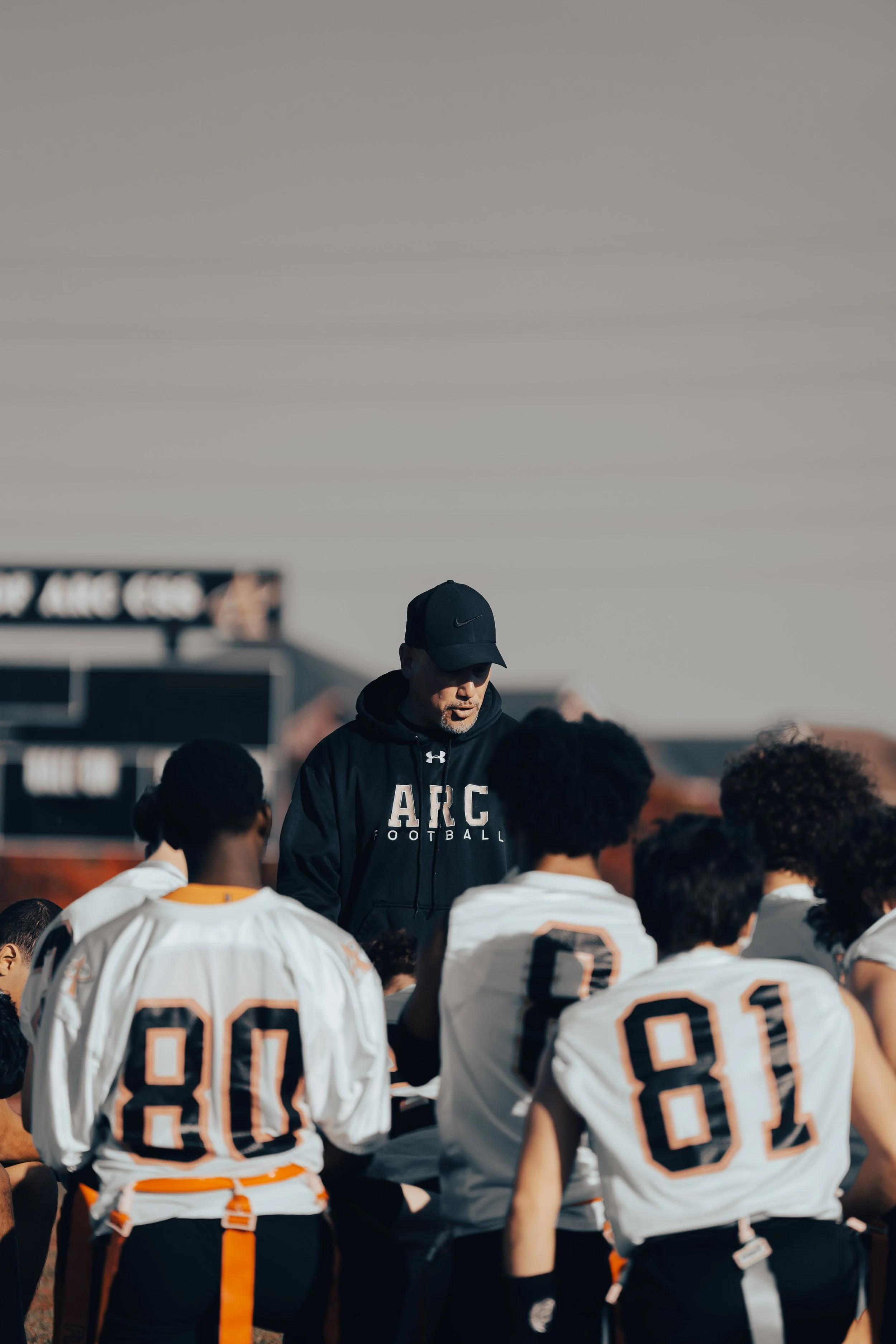 Young football players in white jerseys with black and orange accents, gathered around their coach on a field during daytime.