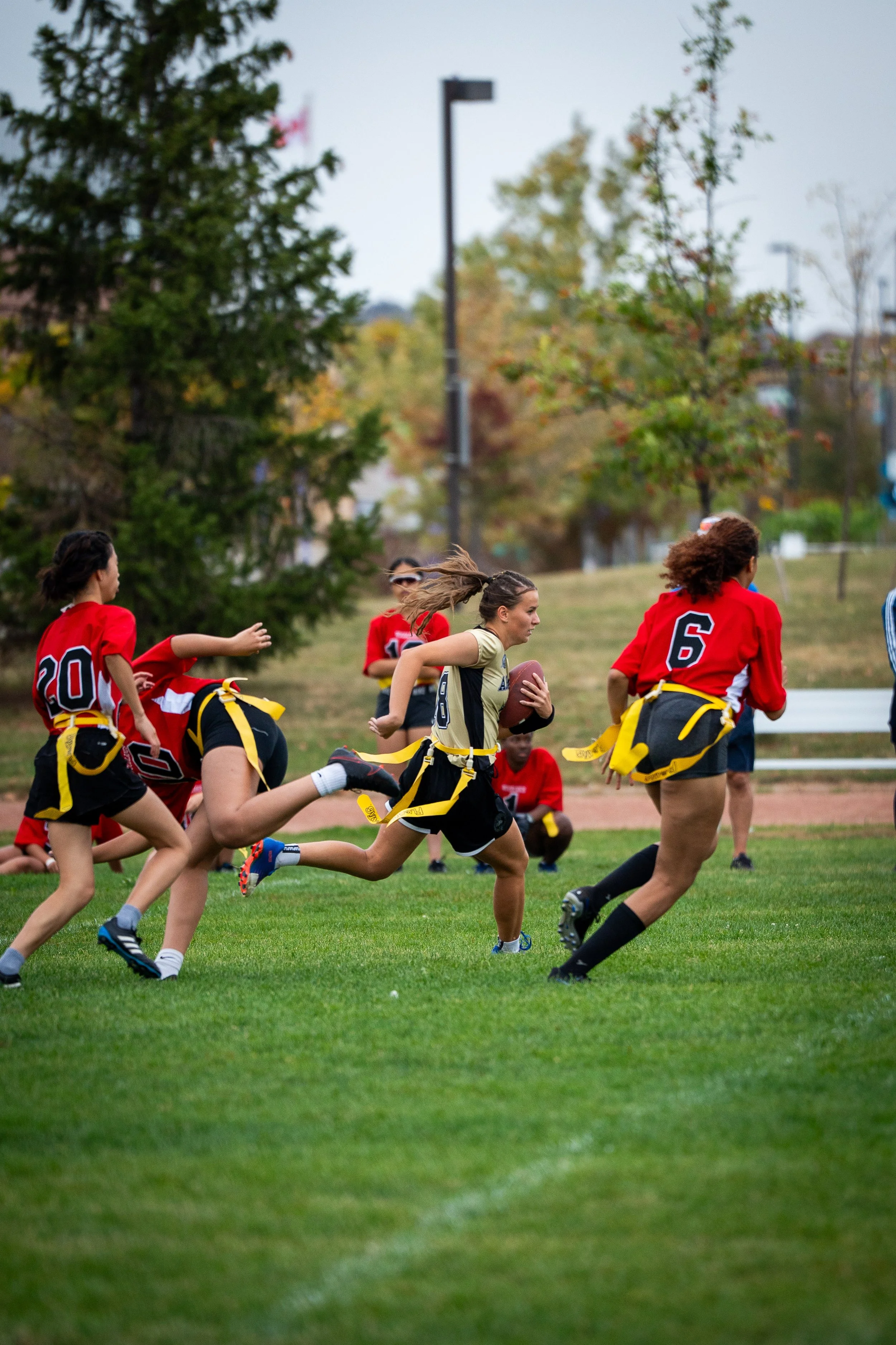 Youth girls playing flag football on a grassy field during daytime, with some players chasing after a girl holding a football.