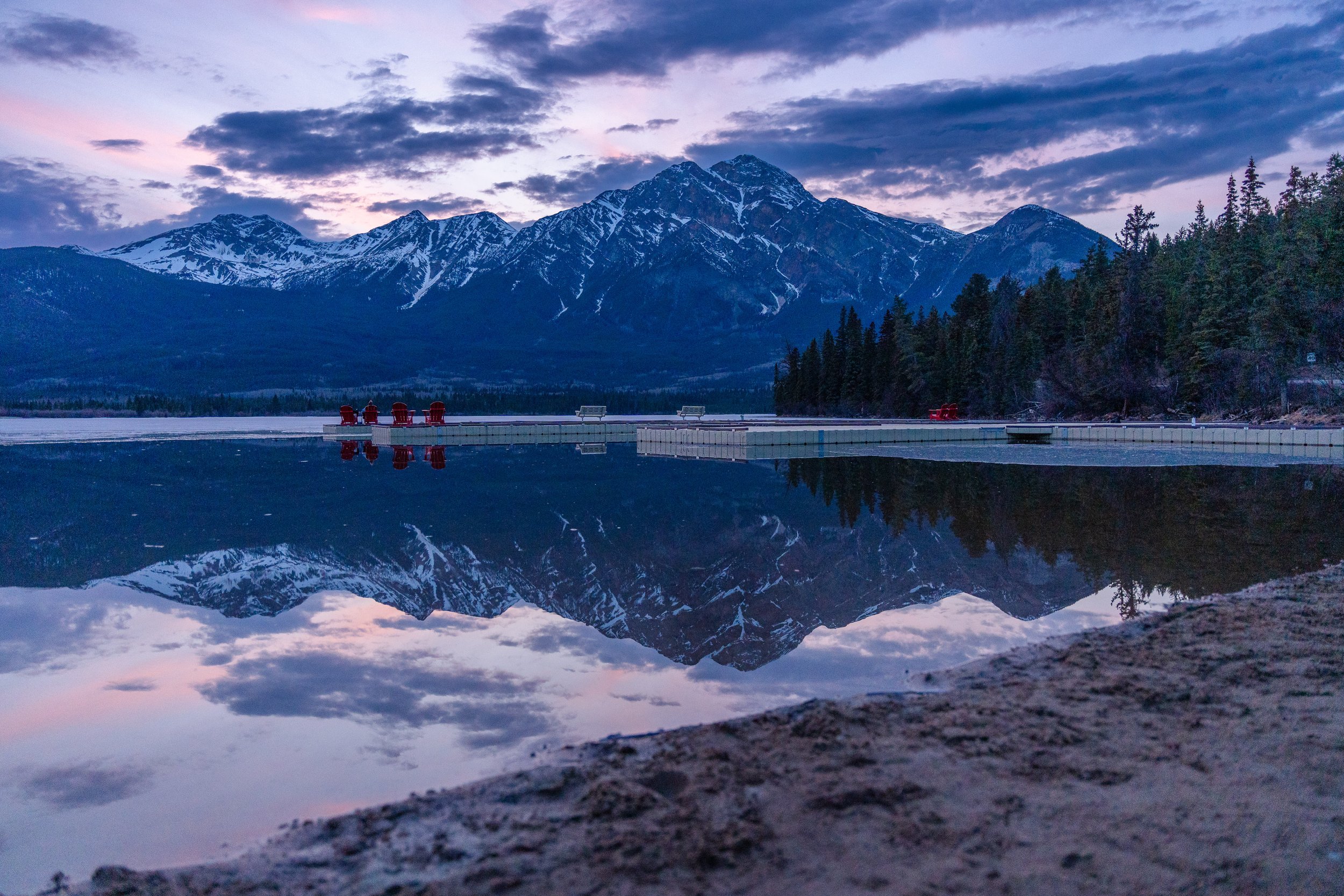 Snow-capped mountains at sunset reflected in a calm lake with a stone edge in the foreground, a dock with red chairs on it, and evergreen trees along the shoreline.